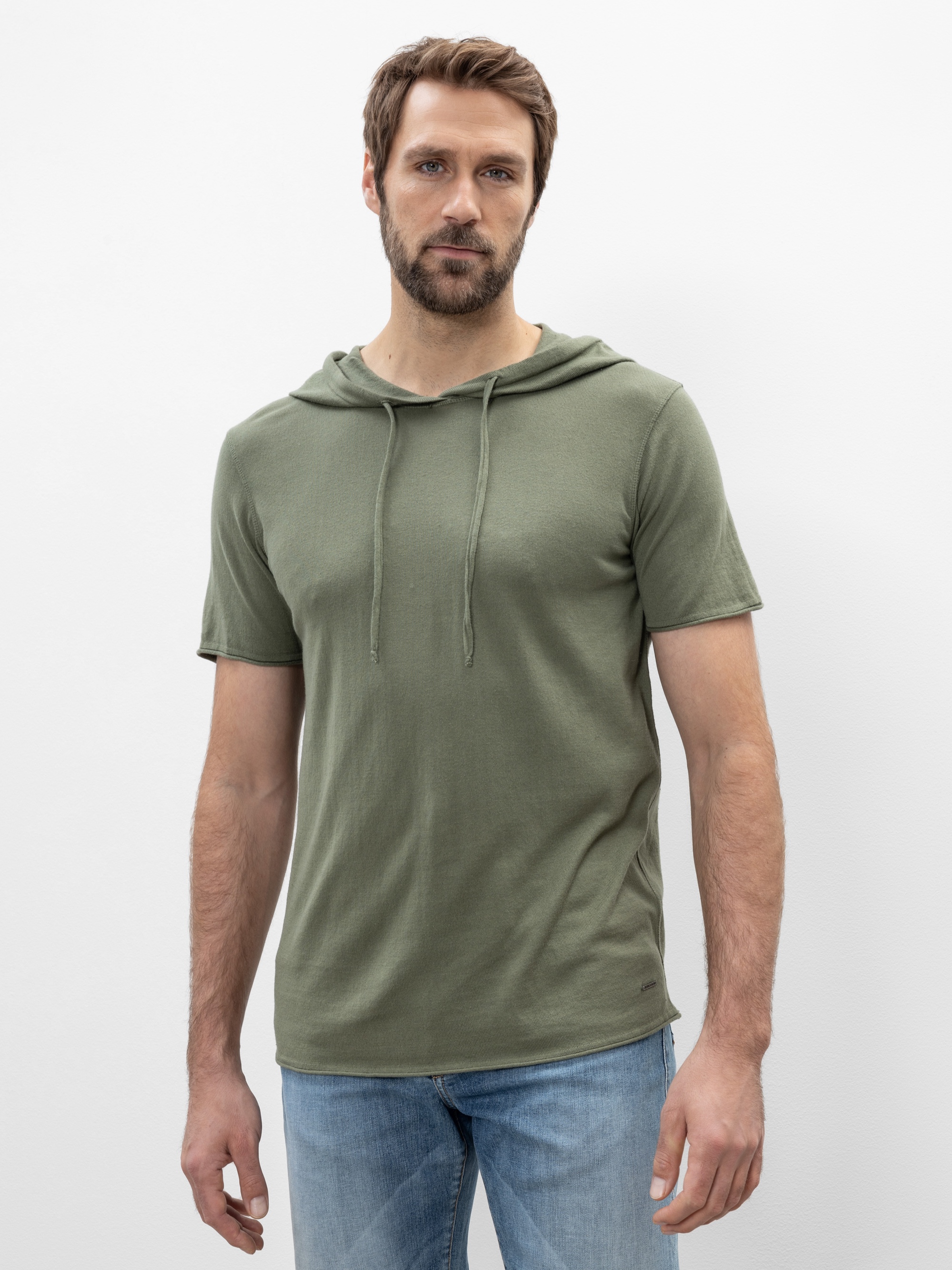 Ein Mann mit kurzen braunen Haaren und einem Bart trägt ein hellolivgrünes, kurzärmeliges Kapuzenshirt und hellblaue Jeans und steht vor einem schlichten weißen Hintergrund.