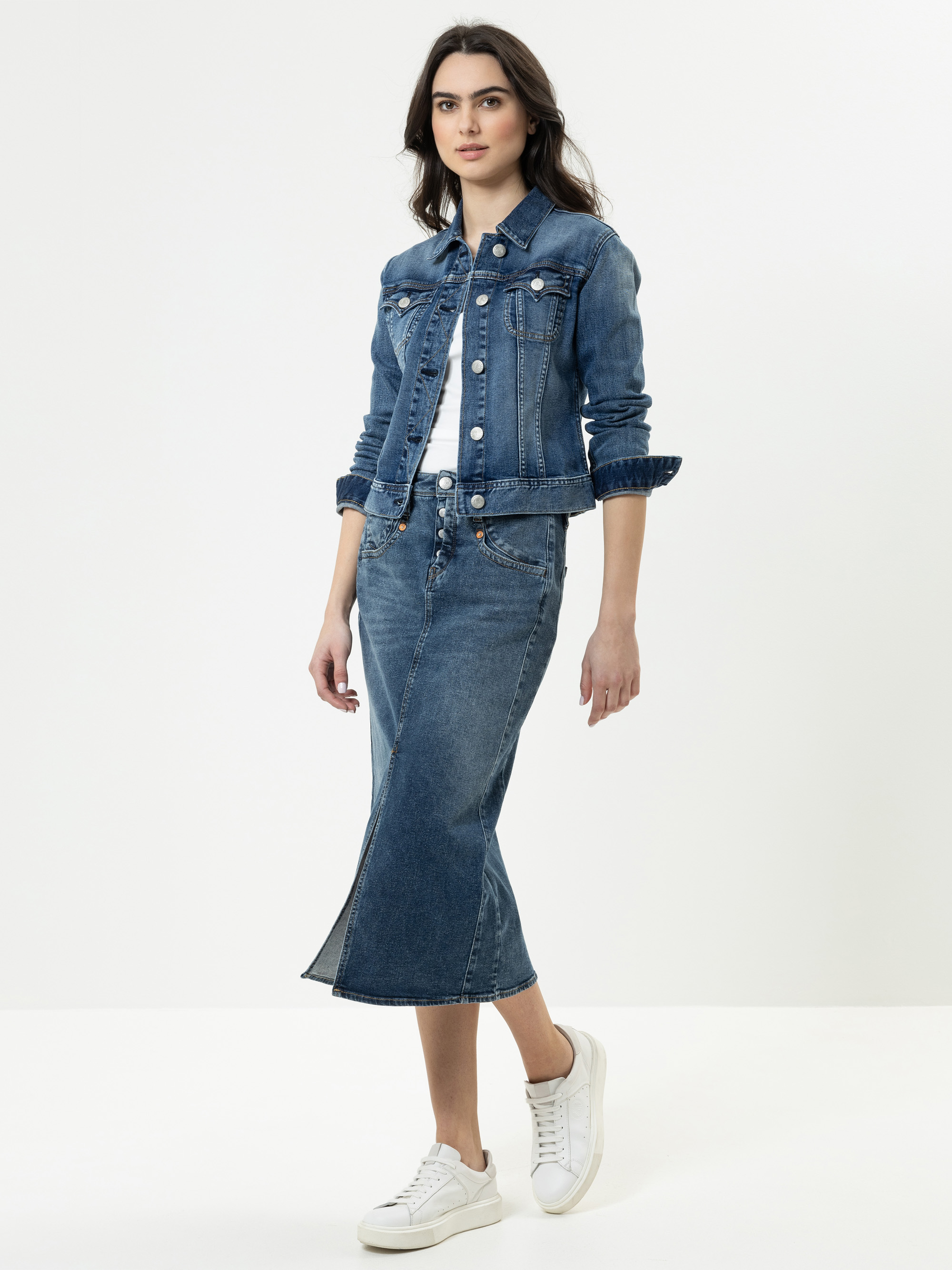 Eine Frau, die eine blaue Jeansjacke und einen dazu passenden blauen Jeans-Midirock trägt, dazu ein weißes Oberteil und weiße Turnschuhe, steht vor einem schlichten weißen Hintergrund.