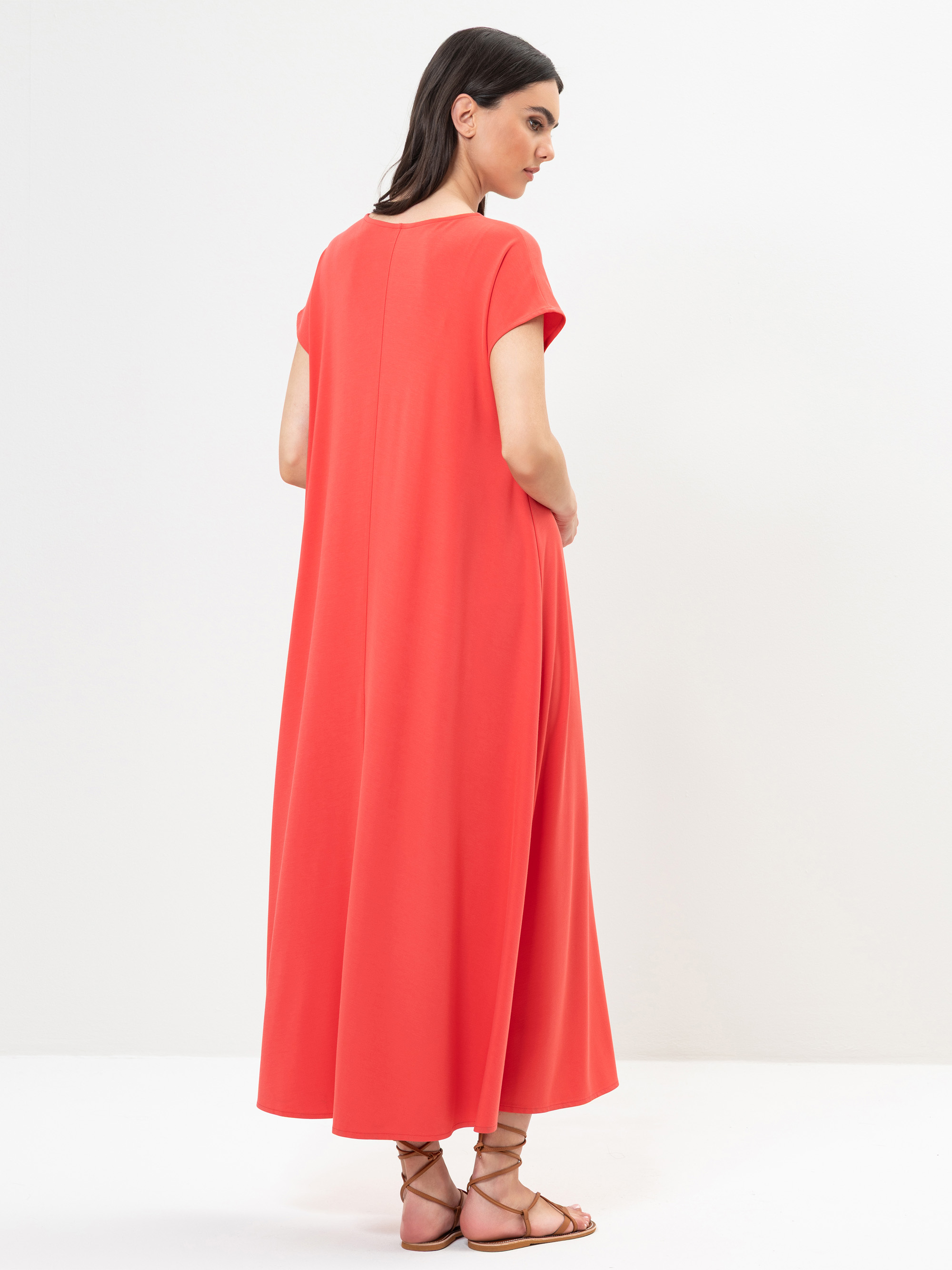 Eine Frau steht in einer minimalistischen Umgebung und trägt ein locker sitzendes, knöchellanges, kurzärmeliges korallenrotes Kleid und braune Riemchensandalen. Sie steht mit dem Rücken zur Kamera und blickt über ihre linke Schulter.