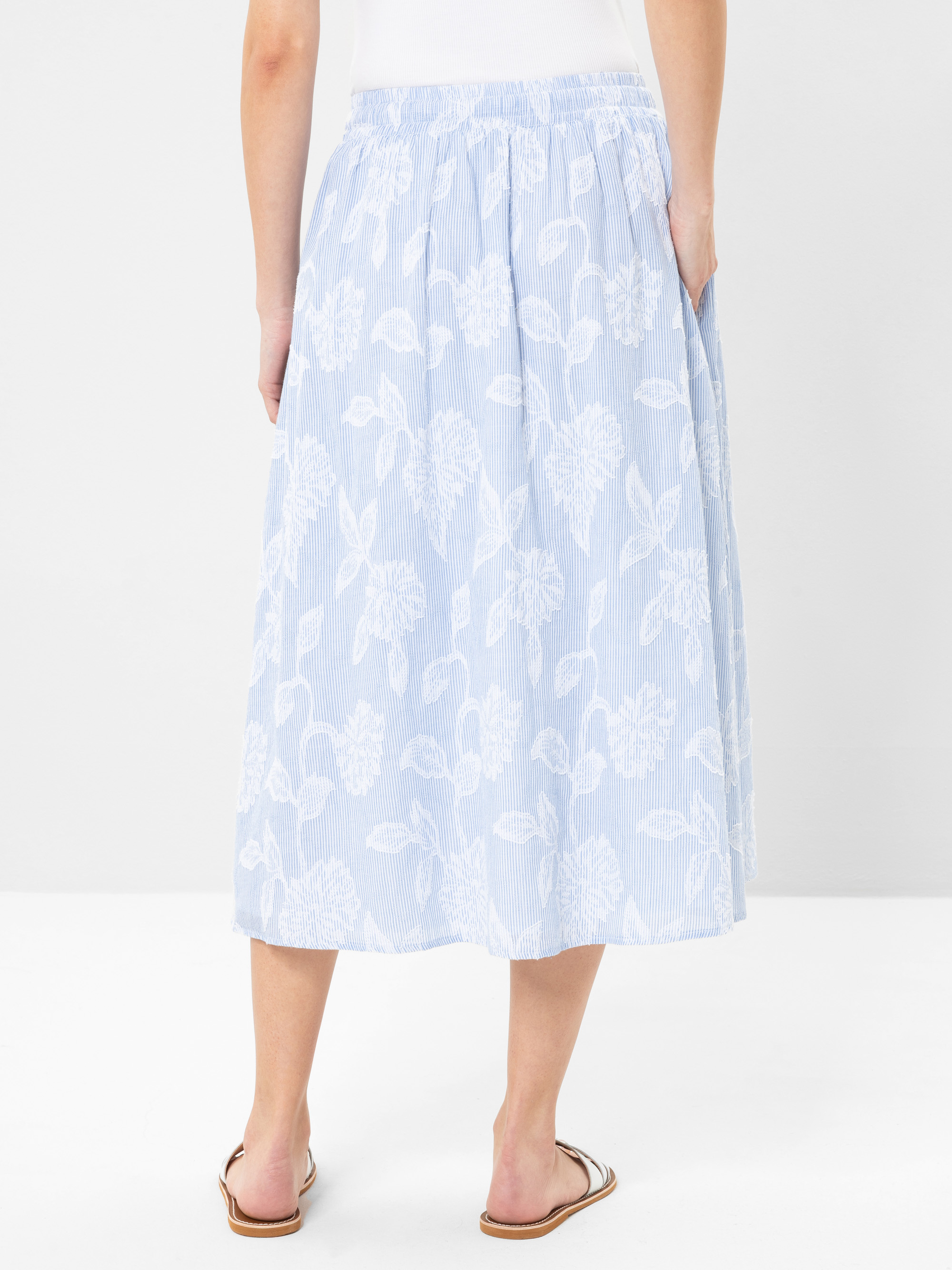 Eine Person, die einen hellblauen, wadenlangen Rock mit weißem Blumenmuster, ein weißes Oberteil und braune Sandalen trägt, steht vor einem schlichten weißen Hintergrund. Das Bild zeigt das Outfit von hinten.