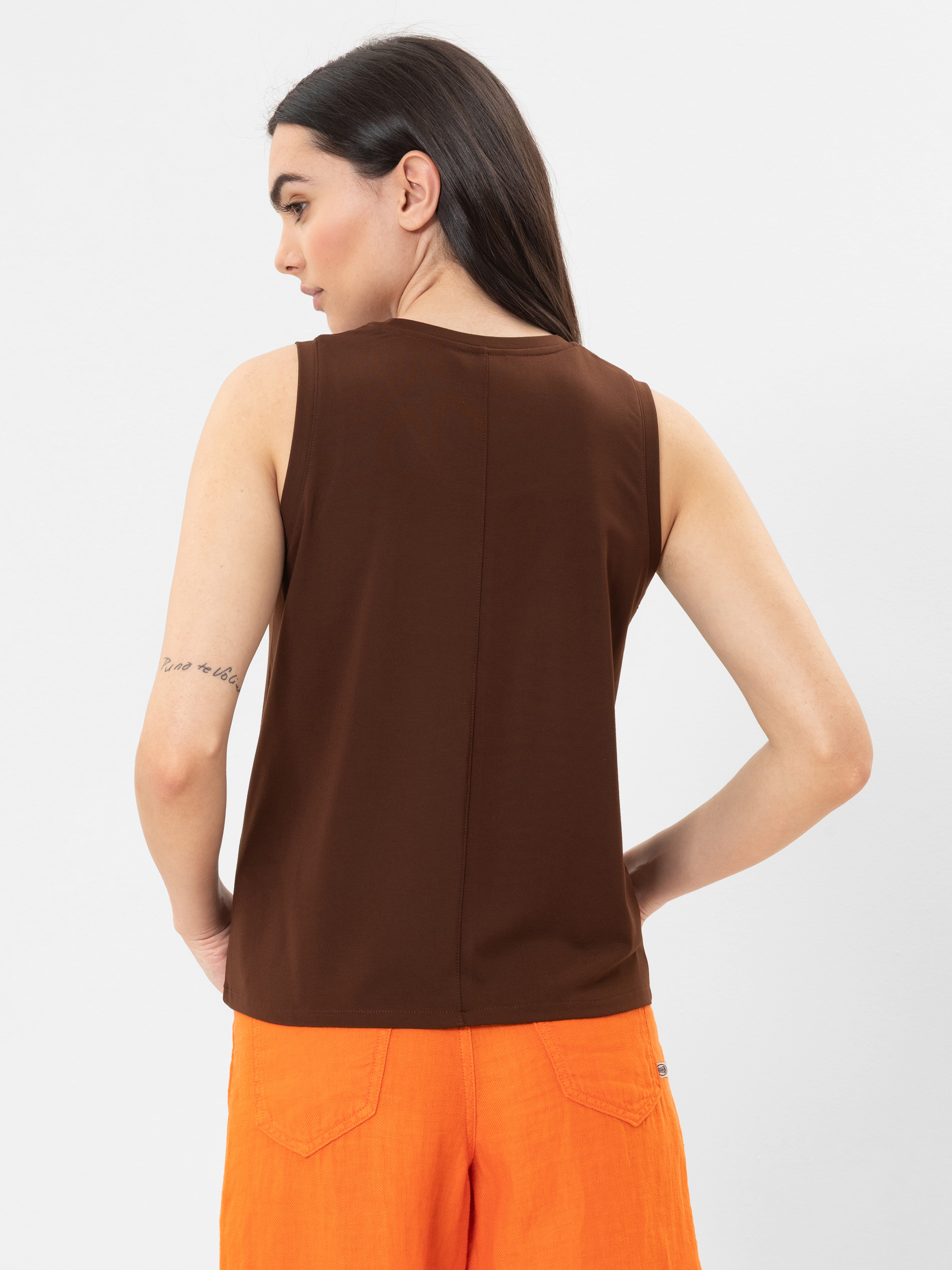 Eine Person mit langen dunklen Haaren ist von hinten zu sehen. Sie trägt ein ärmelloses braunes Oberteil und leuchtend orangefarbene Shorts. Ihr linker Arm hat eine kleine Tätowierung, und der Hintergrund ist einfarbig weiß.