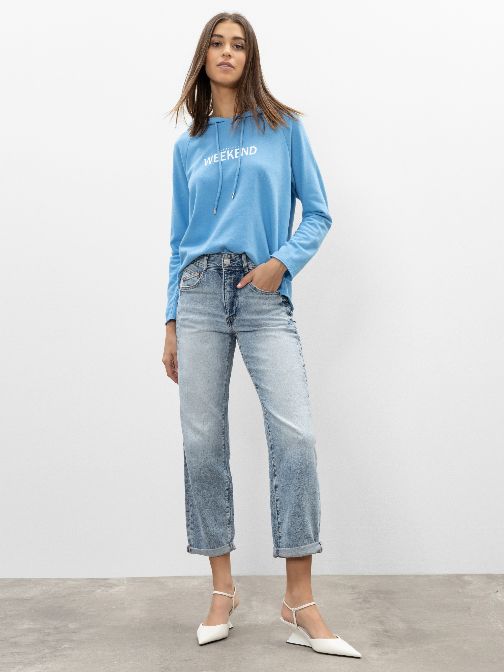 Eine Frau steht an einer schlichten Wand und trägt ein hellblaues, langärmeliges Hemd mit der Aufschrift WEEKEND, helle Jeans mit geradem Bein und weiße Schuhe mit spitzen Zehen.