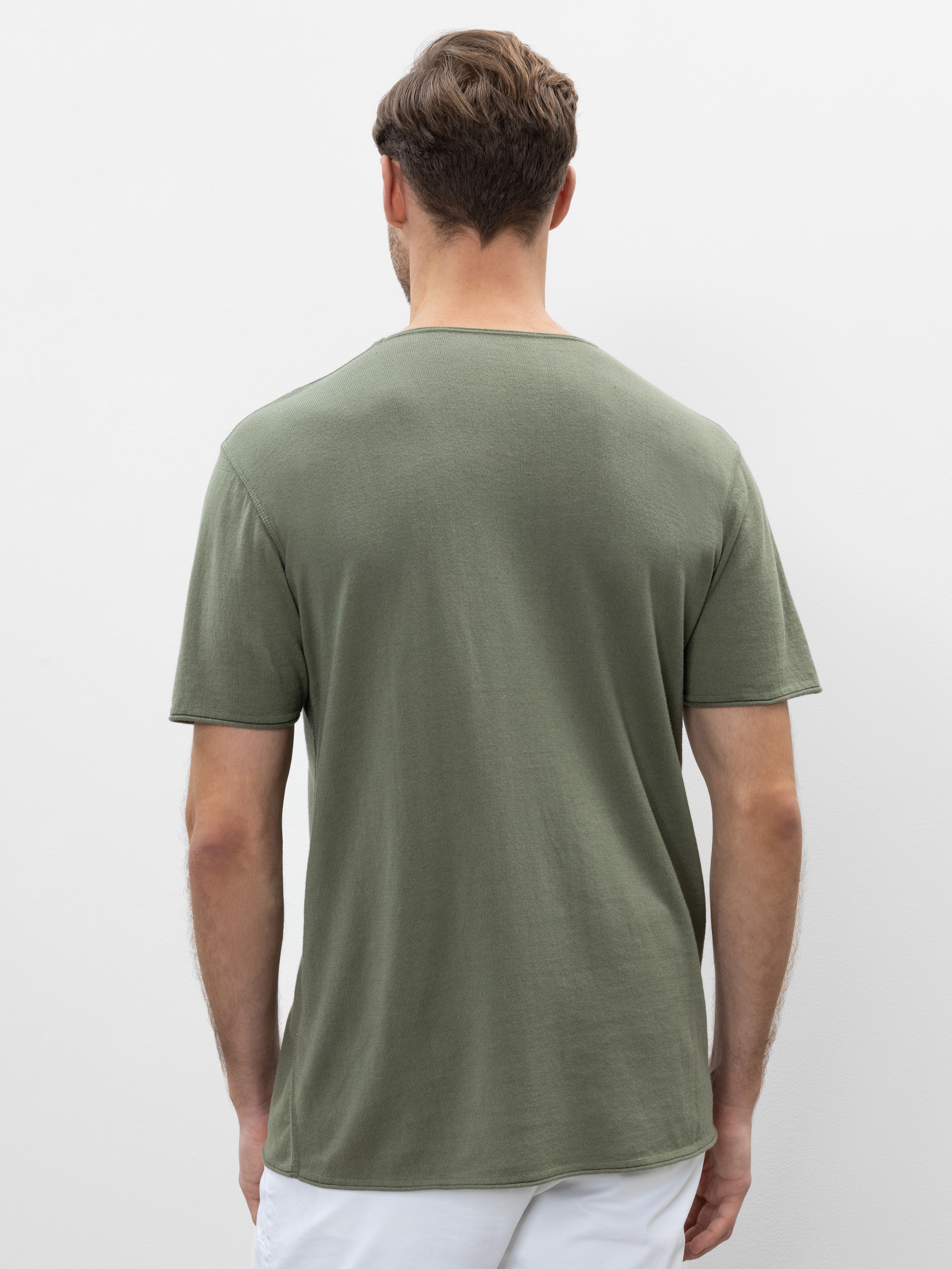 Ein Mann mit kurzen braunen Haaren steht mit dem Rücken zur Kamera, trägt ein olivgrünes, kurzärmeliges T-Shirt und eine weiße Hose vor einem schlichten weißen Hintergrund.