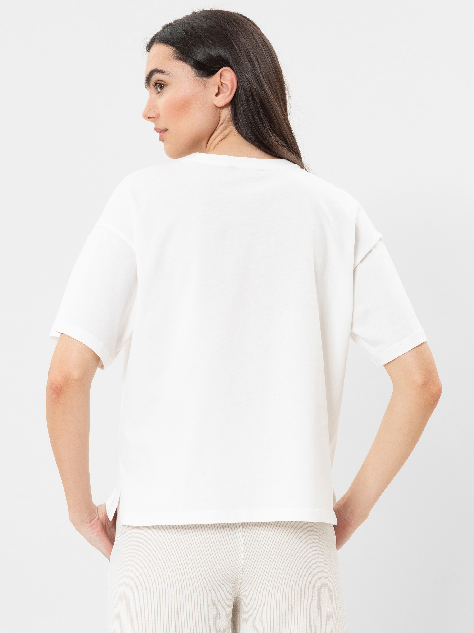 Eine Frau mit langen dunklen Haaren steht mit dem Rücken zur Kamera. Sie trägt ein locker sitzendes weißes T-Shirt und eine helle Hose vor einem schlichten weißen Hintergrund.