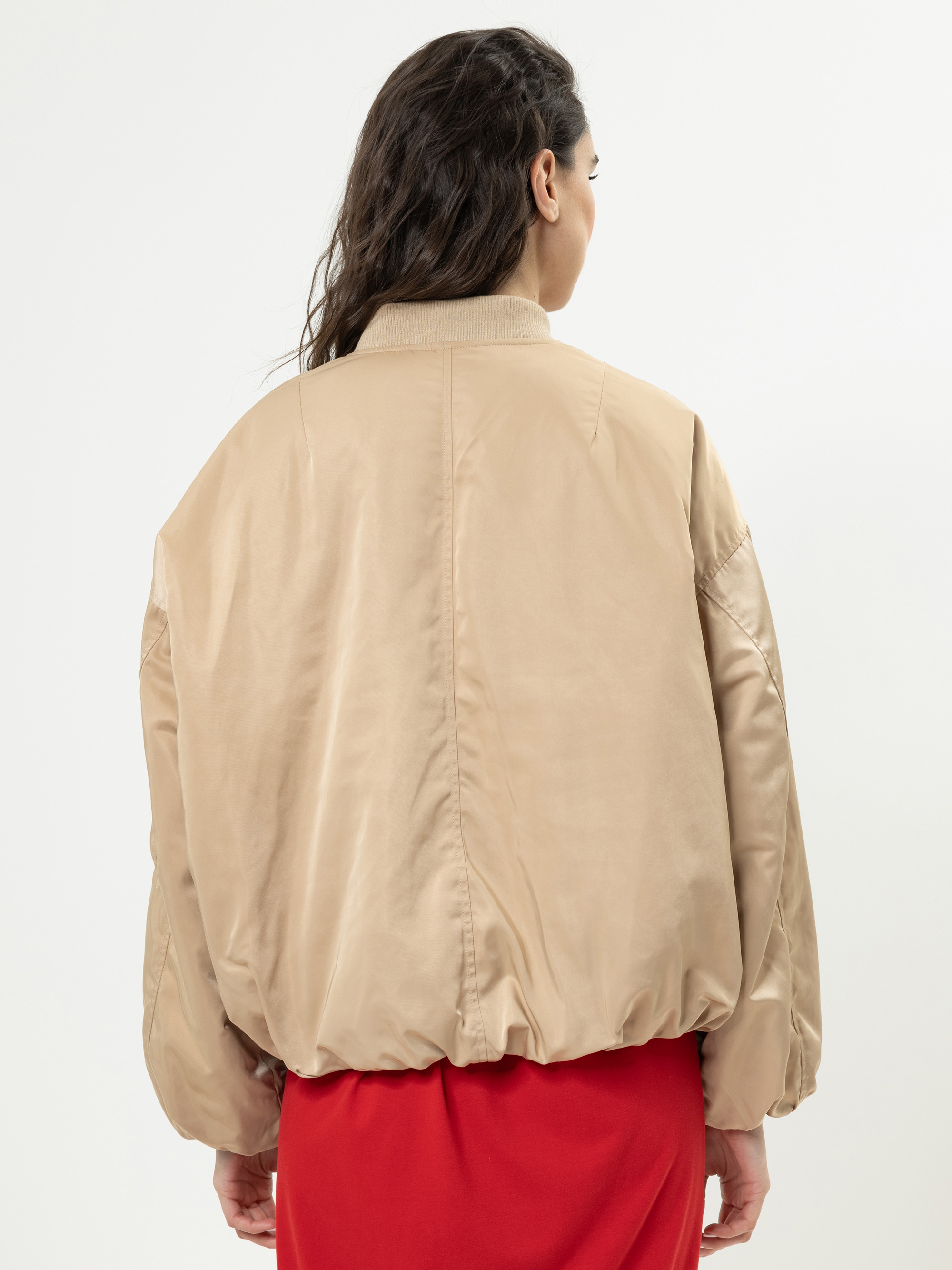 Eine Frau mit gewelltem braunem Haar ist von hinten zu sehen. Sie trägt eine locker sitzende beigefarbene Bomberjacke und einen roten Rock vor einem schlichten weißen Hintergrund.