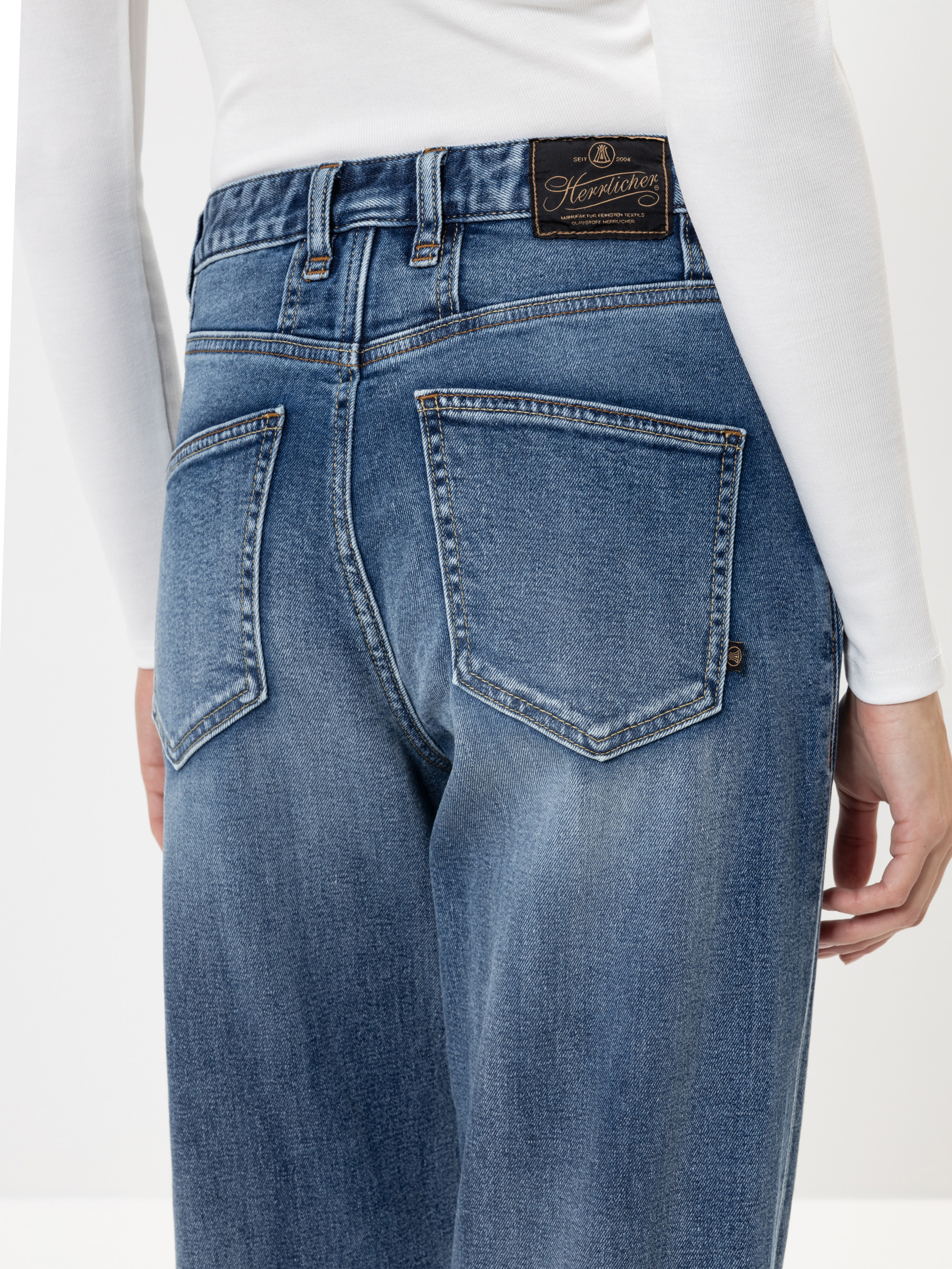 Eine Person trägt eine blaue Denim-Jeans und ein weißes langärmeliges Oberteil, von hinten gesehen. Die Jeans hat Gesäßtaschen und einen schwarzen Markenaufnäher aus Leder am Bund.