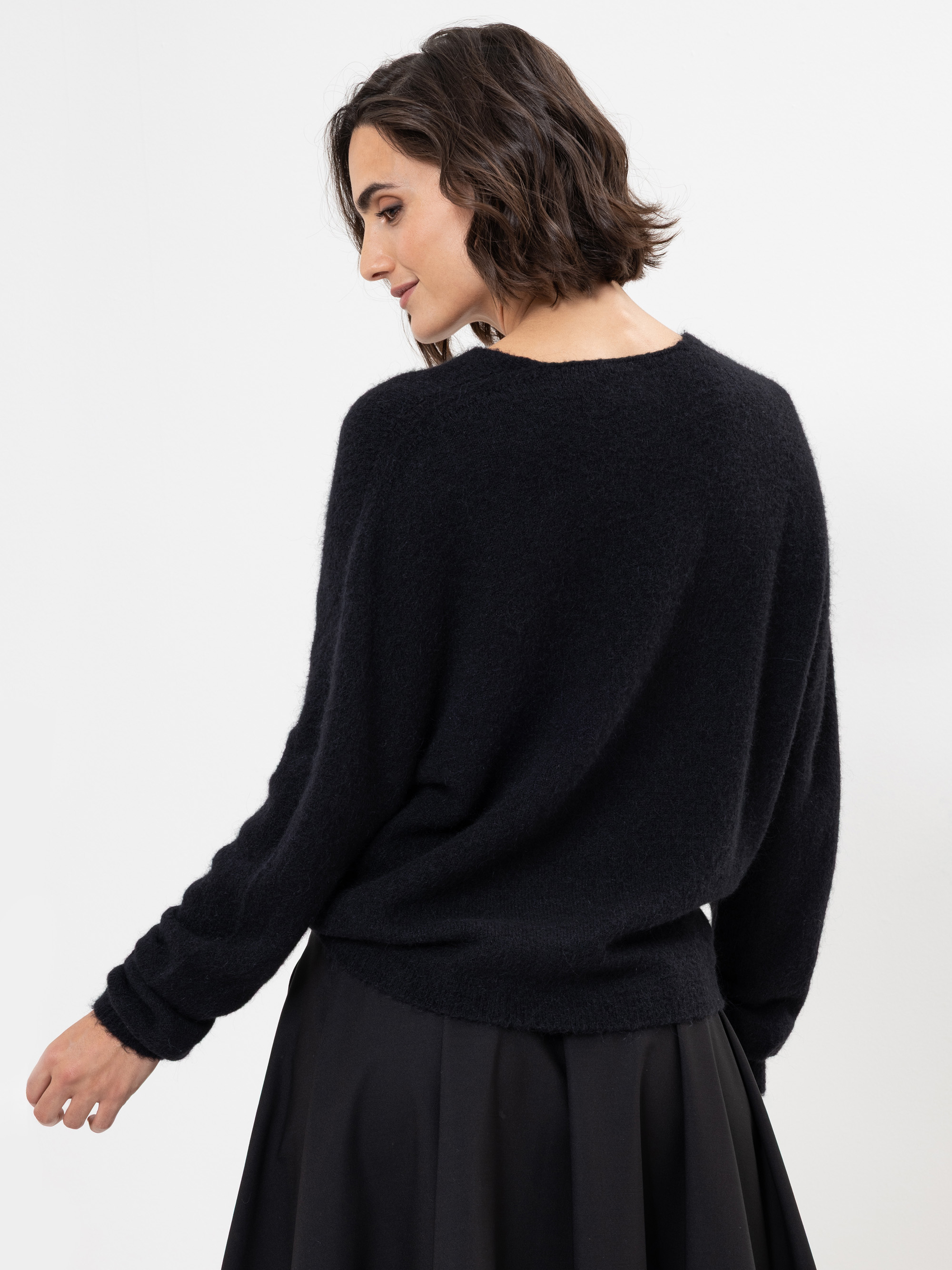 Eine Frau mit kurzen braunen Haaren, die einen lockeren schwarzen Pullover und einen schwarzen Rock trägt, steht leicht von der Kamera abgewandt vor einem schlichten weißen Hintergrund.