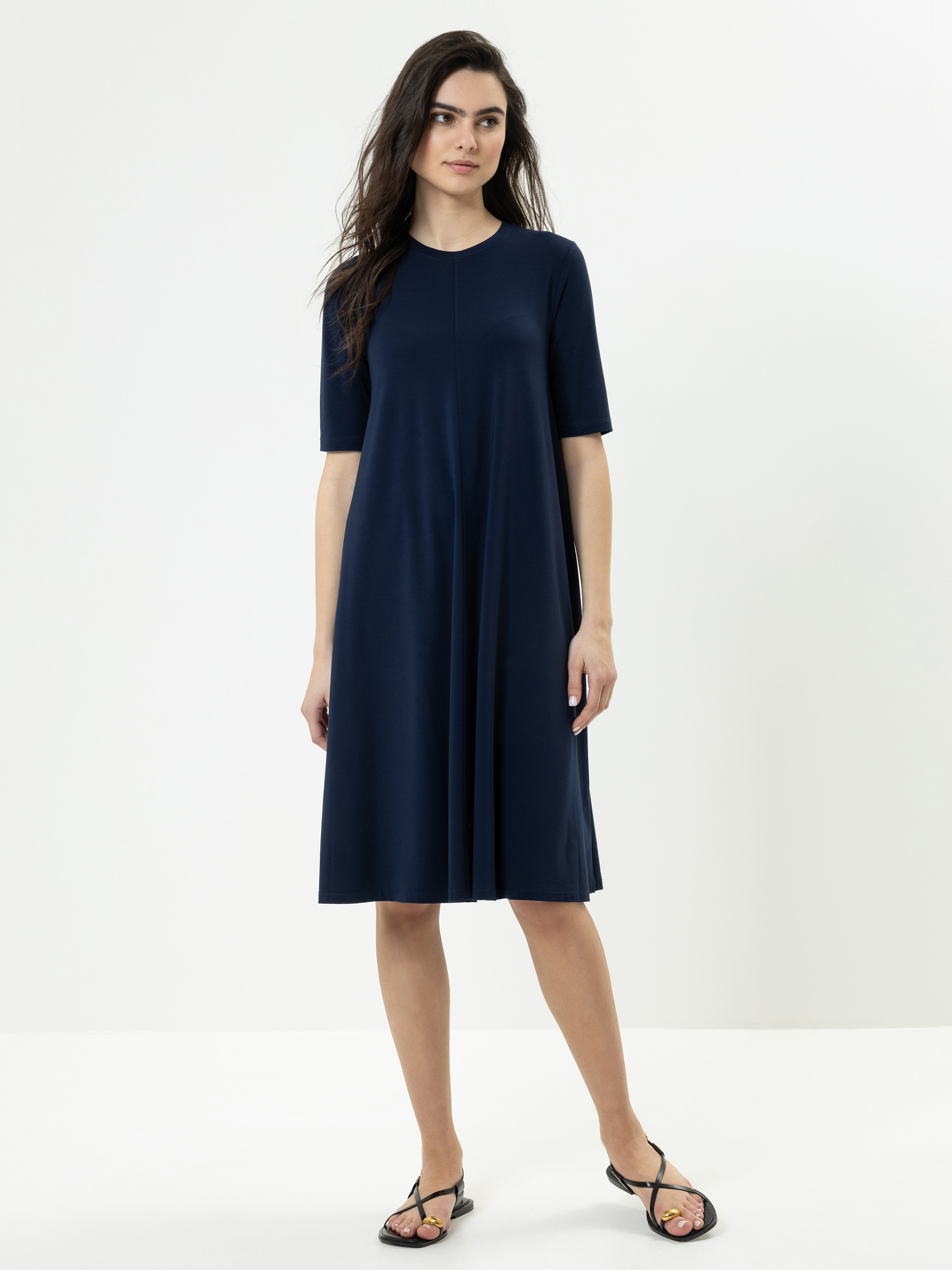 Frau in einem knielangen, marineblauen, kurzärmeligen Kleid mit einfacher, lockerer Passform. Sie hat langes dunkles Haar, steht vor einem schlichten weißen Hintergrund und trägt schwarze Sandalen.