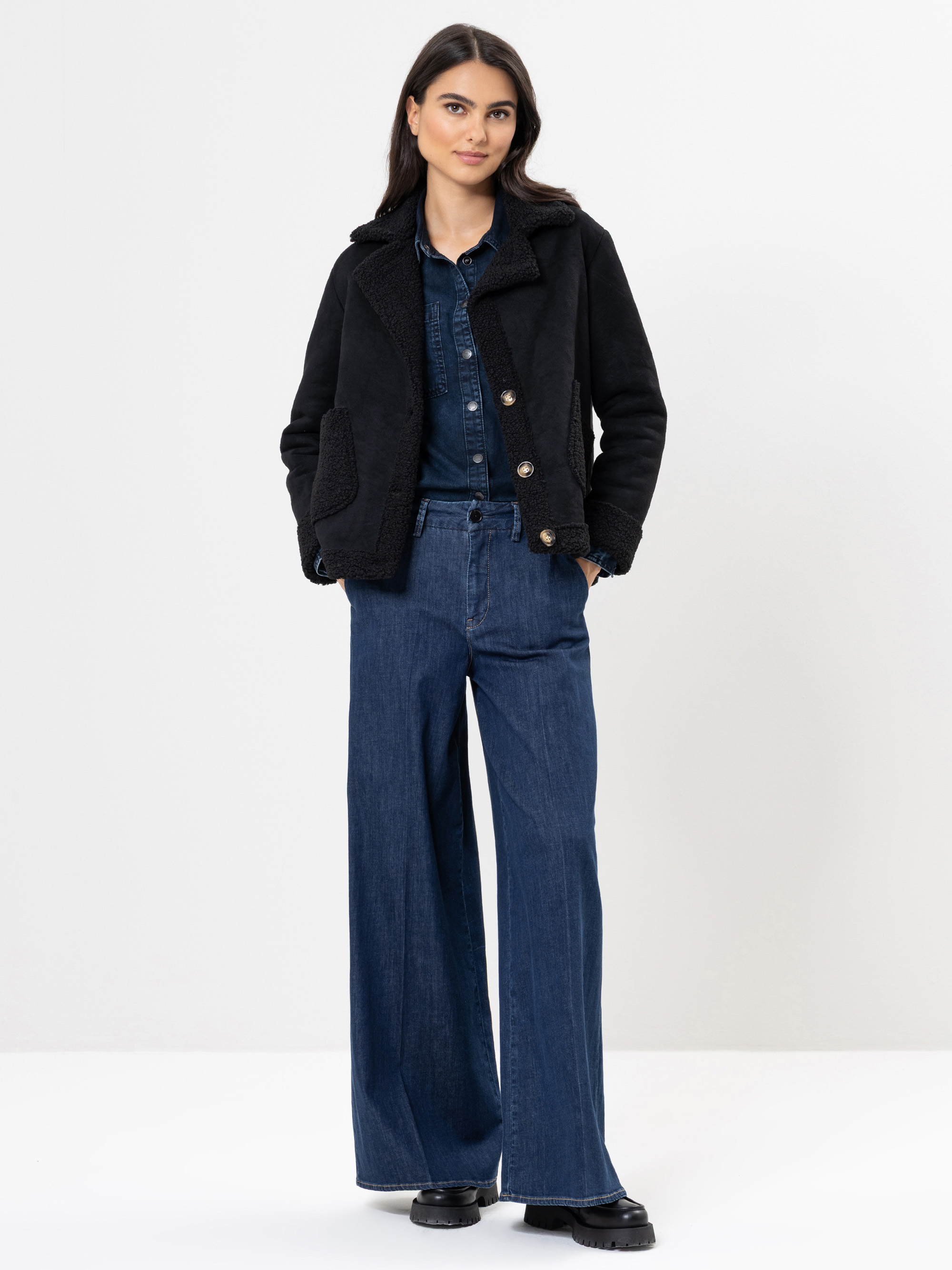 Eine Frau steht vor einem schlichten Hintergrund und trägt eine schwarze Jacke, ein blaues Jeanshemd und eine weit geschnittene blaue Jeans mit schwarzen Schuhen. Sie hat langes dunkles Haar und einen neutralen Gesichtsausdruck.