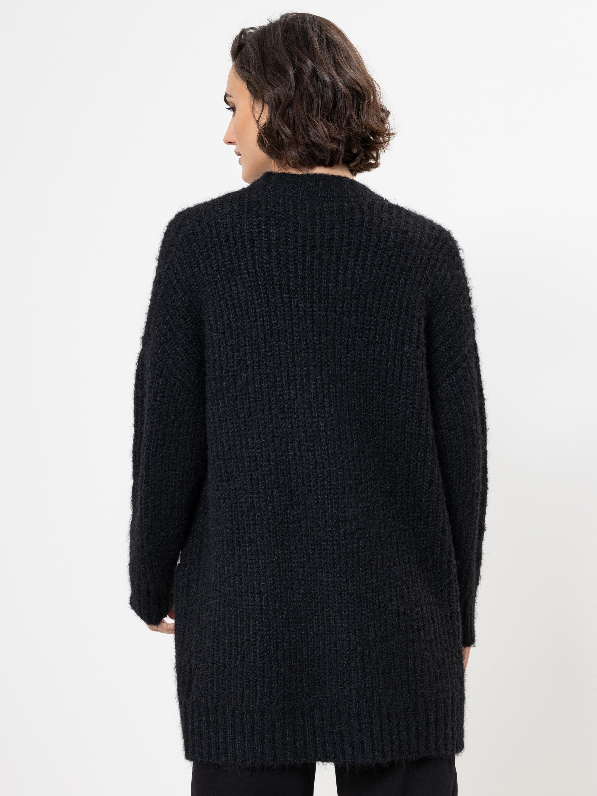 Man sieht eine Person mit kurzem, dunklem Haar von hinten, die einen langen, strukturierten schwarzen Pullover und eine schwarze Hose trägt und vor einem schlichten weißen Hintergrund steht.