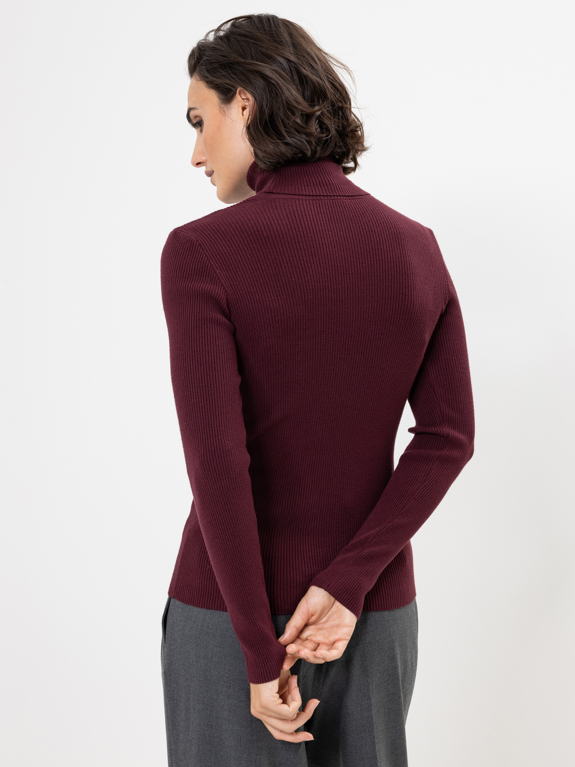 Person, die mit dem Rücken zur Kamera steht, einen langärmeligen, gerippten kastanienbraunen Rollkragenpullover und eine graue Hose trägt und die Hände locker hinter dem Rücken verschränkt. Der Hintergrund ist einfarbig weiß.