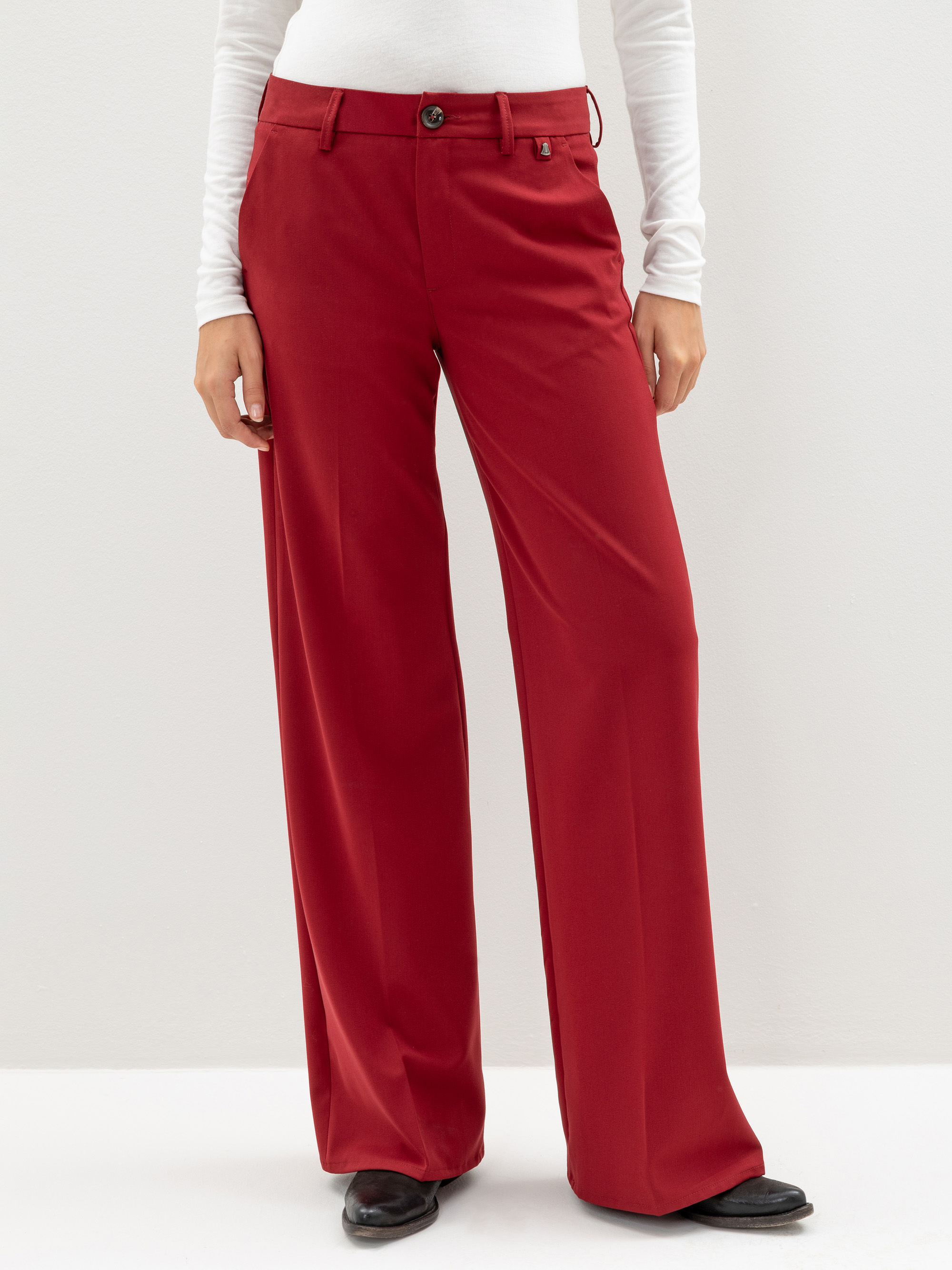 Eine Person, die eine rote Hose mit hoher Taille und weitem Bein sowie ein weißes langärmeliges Oberteil und schwarze Schuhe trägt und vor einem schlichten, hellen Hintergrund steht.