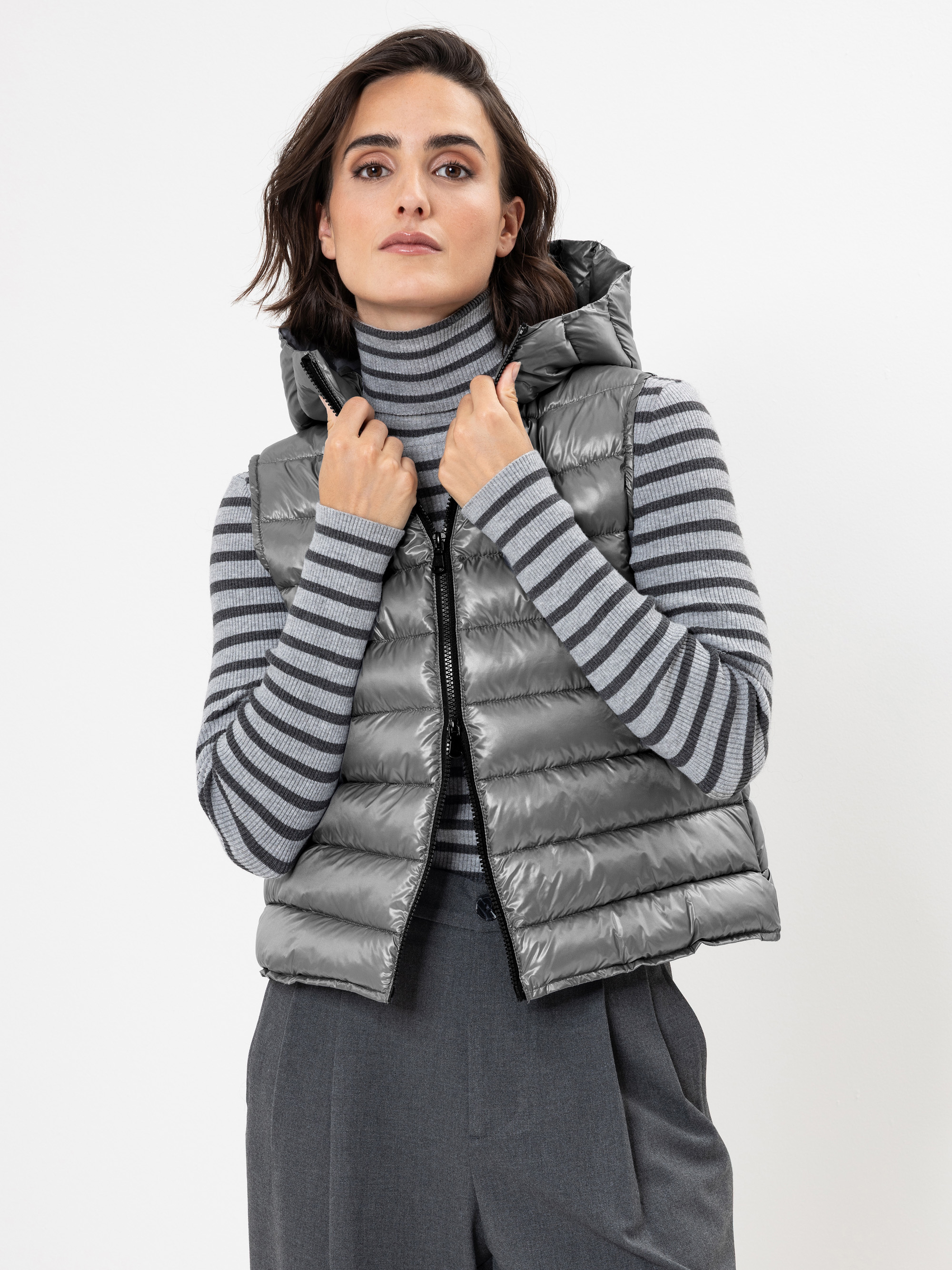 Eine Frau, die eine glänzende graue Pufferweste über einem grau-weiß gestreiften Rollkragenpullover und einer grauen Hose trägt, steht vor einem schlichten weißen Hintergrund und hält den Kragen der Weste mit beiden Händen.
