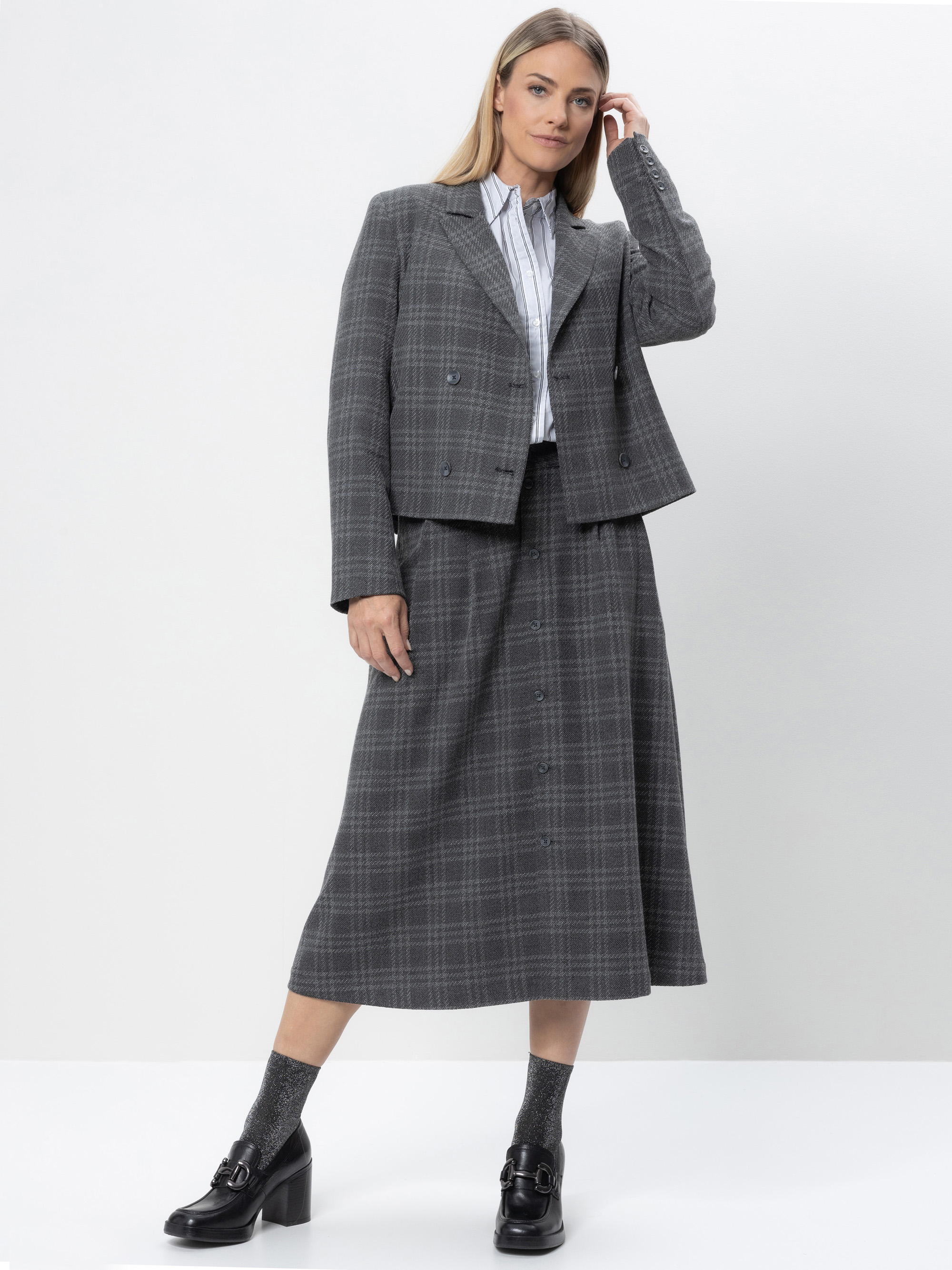 Eine Frau steht vor einem schlichten Hintergrund und trägt einen grau karierten Blazer und einen dazu passenden Rock in A-Linie, eine weiße Bluse mit dünnen Streifen, graue Socken und schwarze Halbschuhe mit Absätzen. Sie hat langes, glattes blondes H