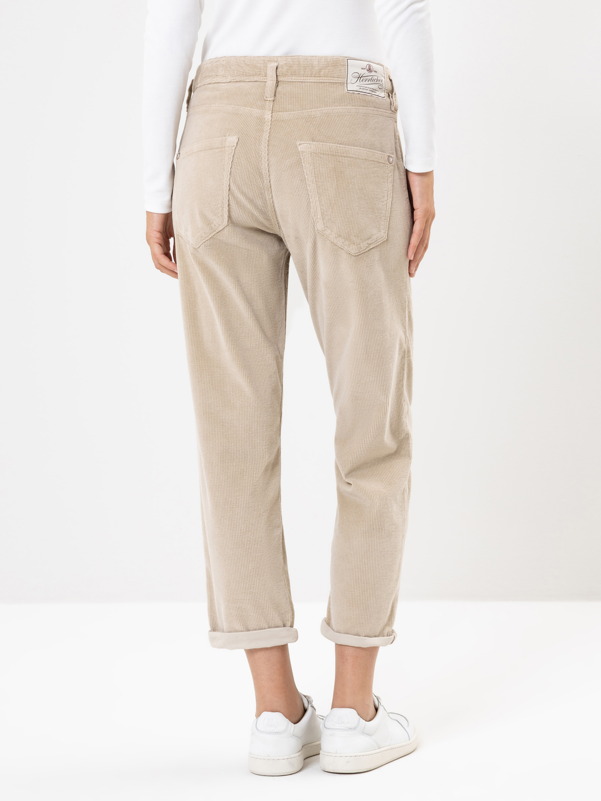 Eine Person steht mit dem Gesicht nach hinten und trägt eine hellbeige Cordhose, ein weißes langärmeliges Oberteil und weiße Turnschuhe. Die Hose ist an den Knöcheln mit Bündchen versehen, und am hinteren Bund ist ein Etikett zu sehen.
