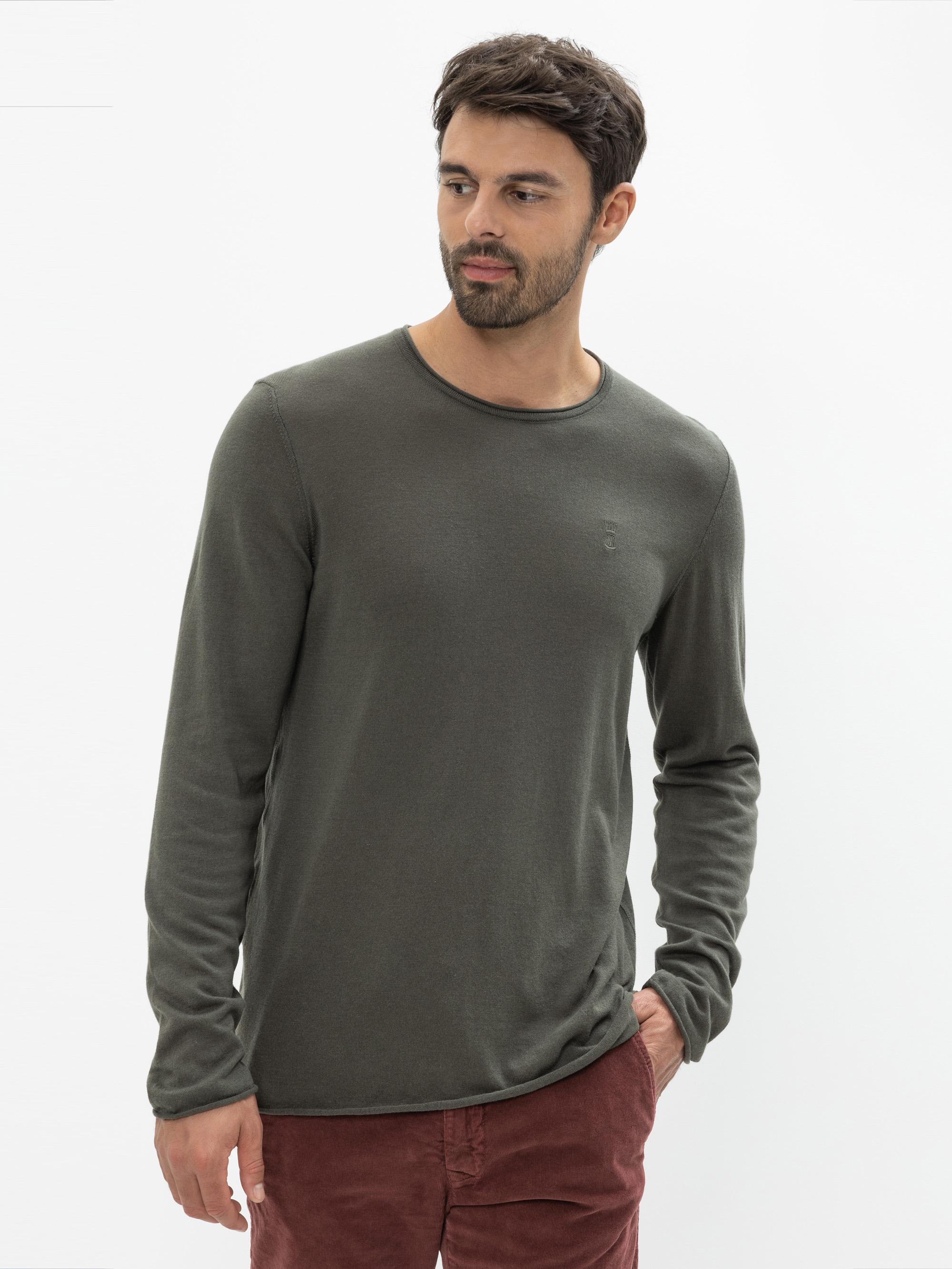 Ein Mann mit kurzen dunklen Haaren und einem Bart trägt ein langärmeliges, olivgrünes Hemd und eine rote Hose. Er steht vor einem schlichten weißen Hintergrund, hat eine Hand in der Tasche und schaut nach links.