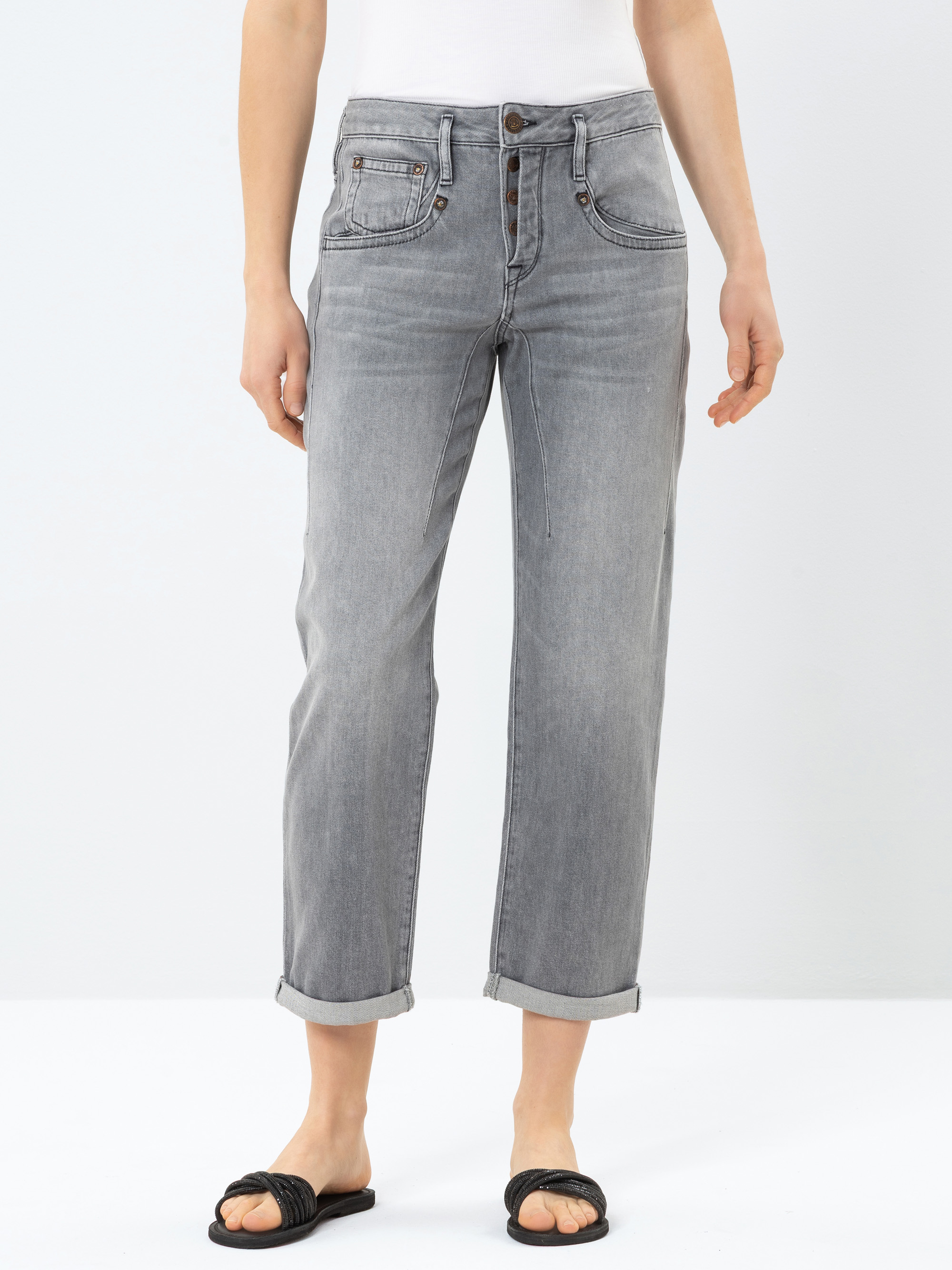 Eine Person, die eine hellgraue, hochgekrempelte Jeans, ein weißes Tanktop und schwarze Slipper trägt, steht vor einem schlichten weißen Hintergrund.