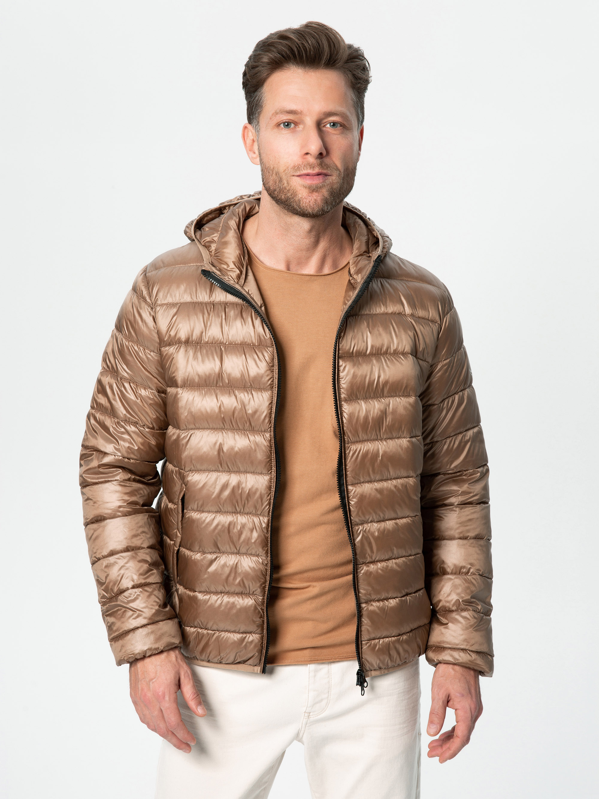 Ein Mann steht vor einem schlichten Hintergrund, trägt eine glänzende hellbraune Pufferjacke über einem hellbraunen Hemd und einer weißen Hose und schaut mit neutralem Blick in die Kamera.
