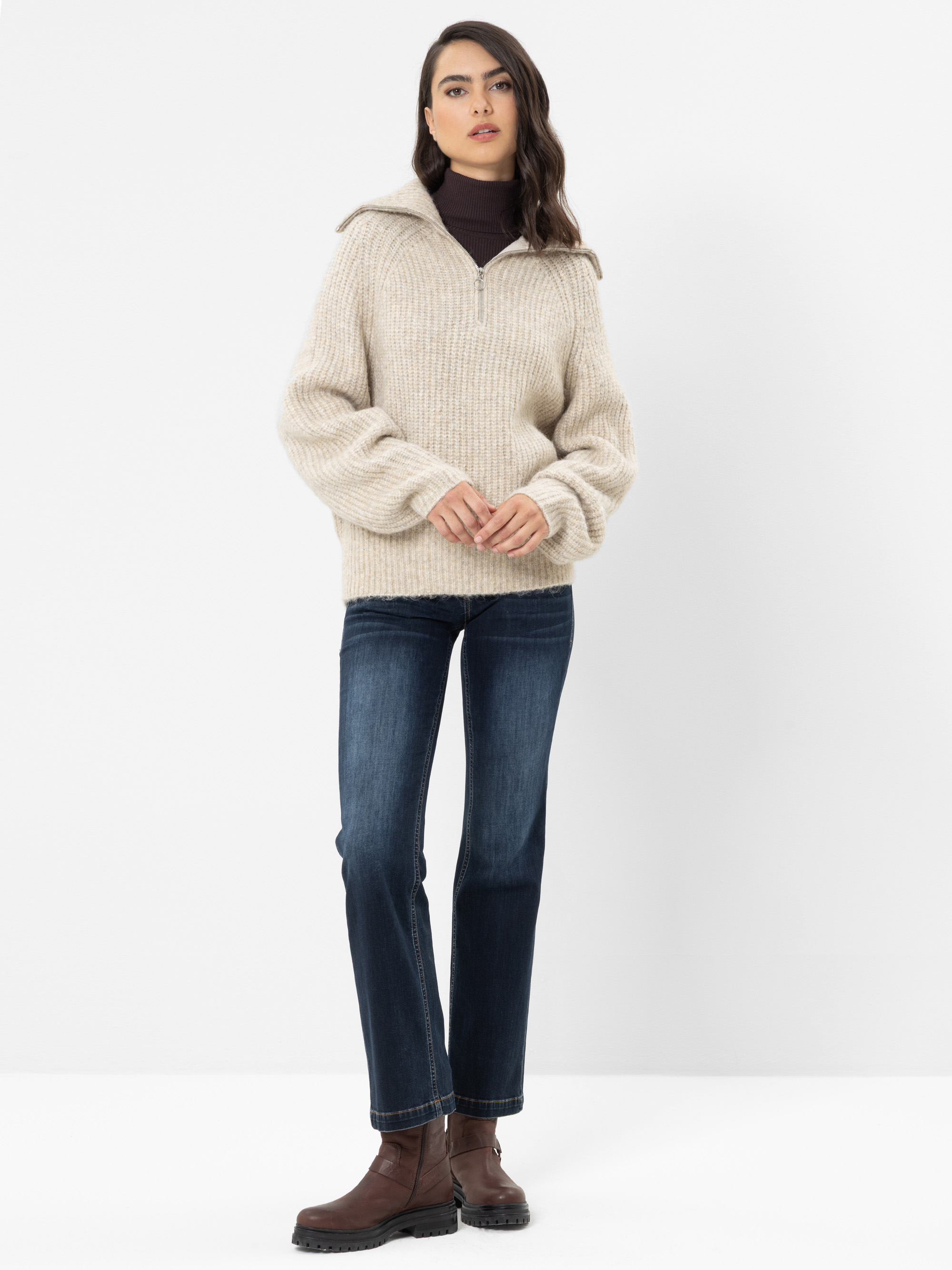 Eine Person steht vor einem weißen Hintergrund und trägt einen beigen Strickpullover, eine dunkle Jeans, braune Stiefel und ein dunkles Unterhemd. Sie hat langes, welliges Haar und einen neutralen Gesichtsausdruck.
