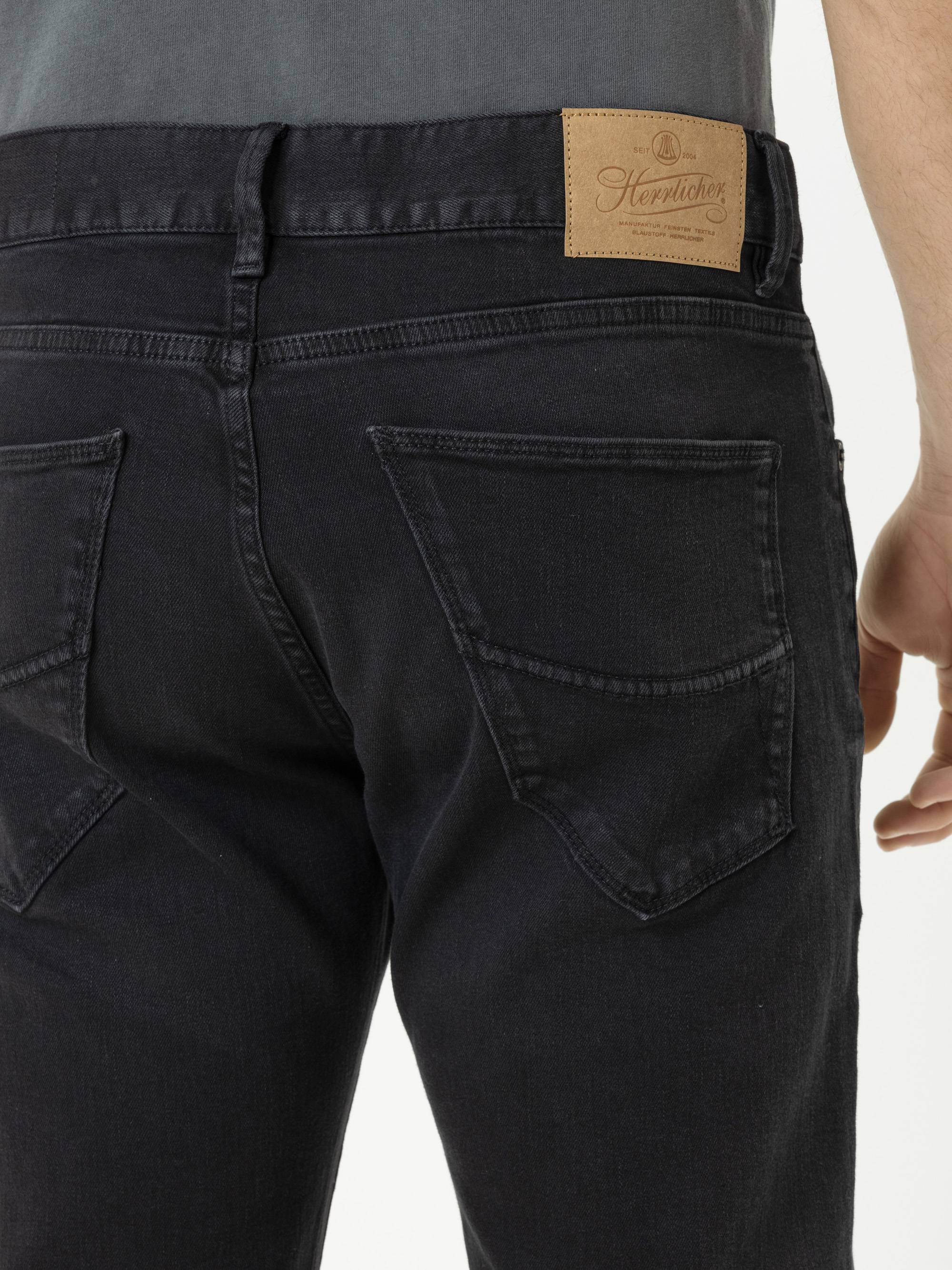 Nahaufnahme des Rückens einer Person, die eine dunkle Jeans und ein graues Hemd trägt, mit zwei Gesäßtaschen und einem braunen Lederaufnäher am Hosenbund.