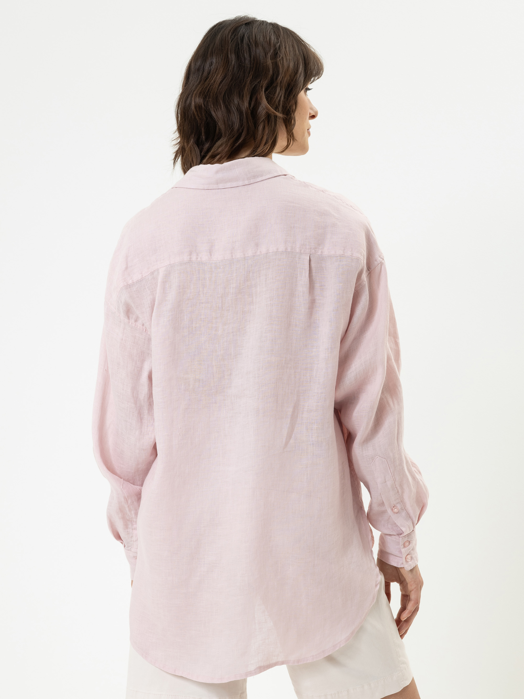 Eine Person mit gewelltem braunem Haar ist von hinten zu sehen. Sie trägt ein locker sitzendes, hellrosa langärmeliges Hemd und weiße Shorts vor einem einfarbig weißen Hintergrund.