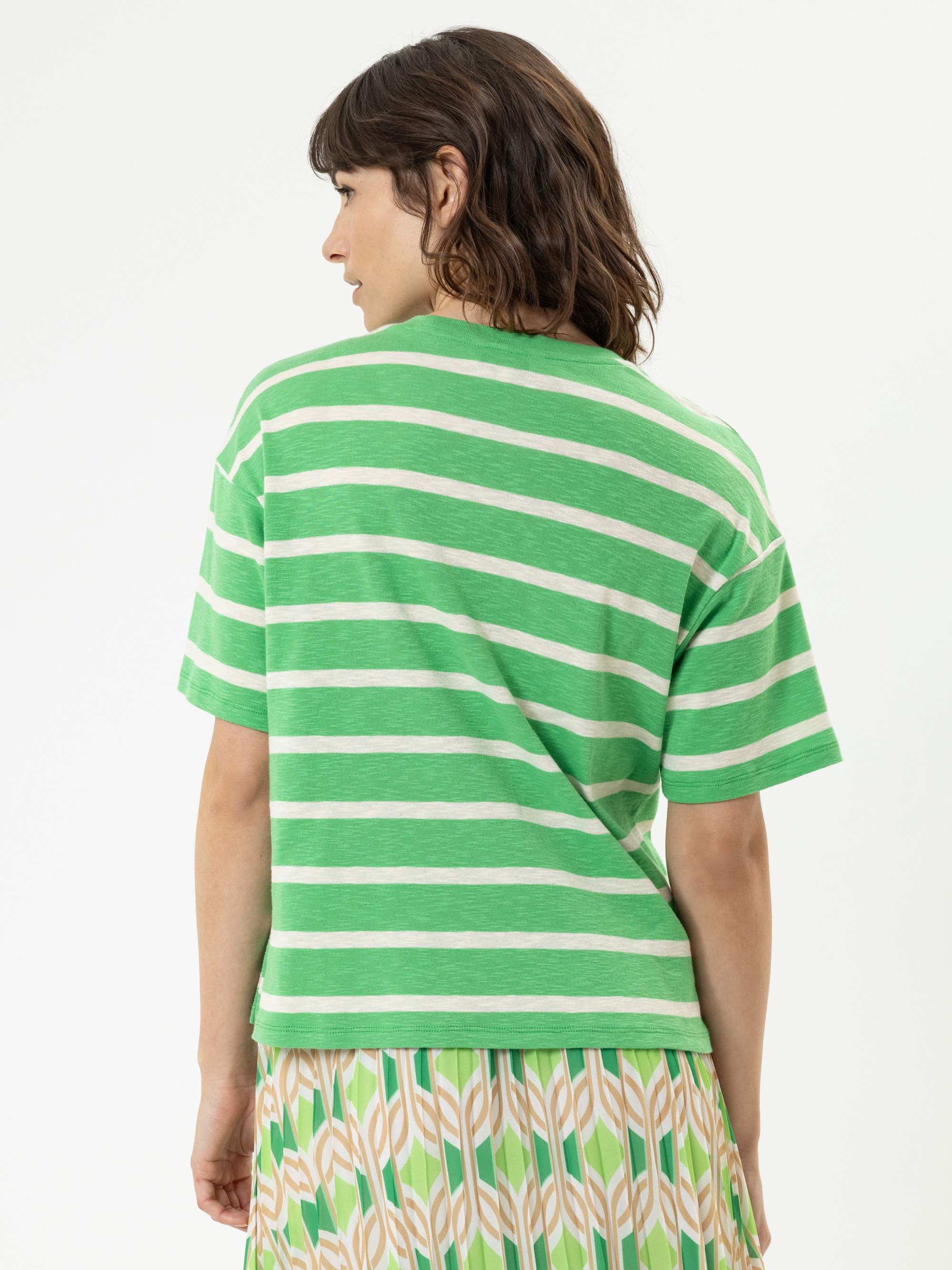 Eine Person mit kurzen braunen Haaren wird von hinten gezeigt. Sie trägt ein grün-weiß gestreiftes kurzärmeliges Hemd und einen gemusterten Rock mit grünen, beigen und orangefarbenen Formen auf weißem Hintergrund.