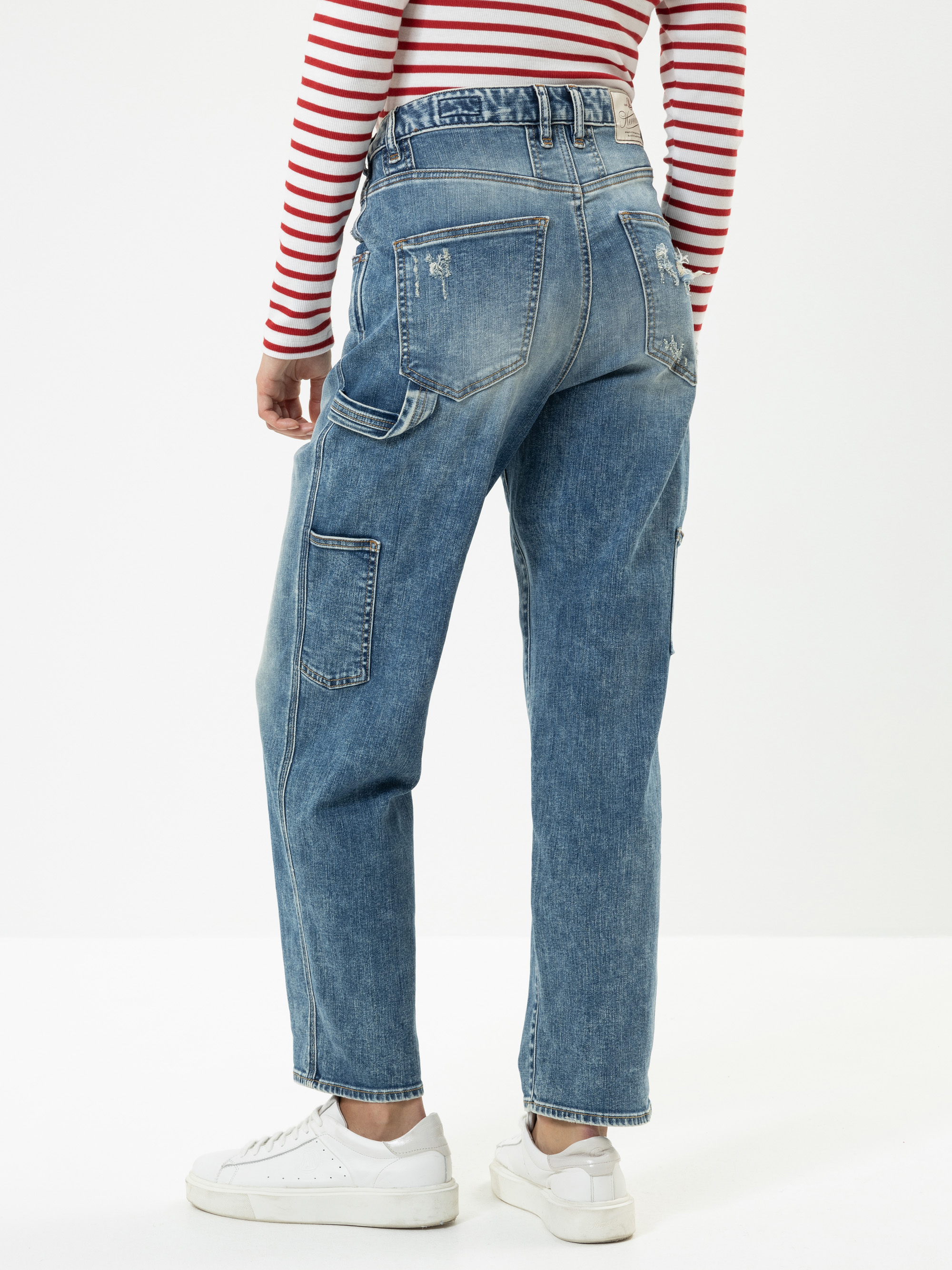 Eine Person, die eine blaue Jeans mit hoher Taille und mehreren Taschen, ein rot-weiß gestreiftes langärmeliges Hemd und weiße Turnschuhe trägt, steht mit dem Gesicht zur Wand vor einem einfarbig weißen Hintergrund.