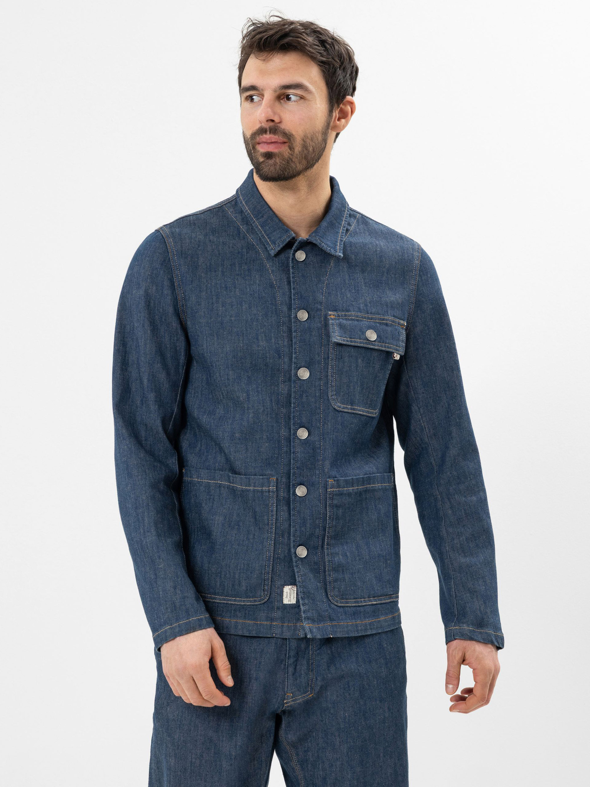 Ein Mann mit Bart, der eine blaue Jeansjacke mit silbernen Knöpfen und eine passende Jeanshose trägt, steht vor einem schlichten weißen Hintergrund.