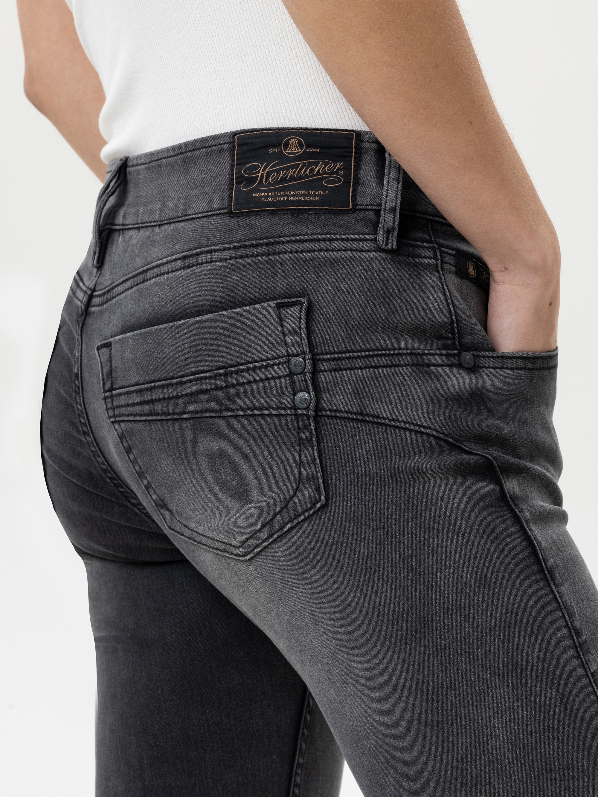 Eine Person, die eine dunkelgraue Jeans und ein weißes geripptes Oberteil trägt, steht mit einer Hand in der Tasche. Die Jeans hat sichtbare Nähte, Gesäßtaschen und ein Lederetikett der Marke Herrlicher am Bund.