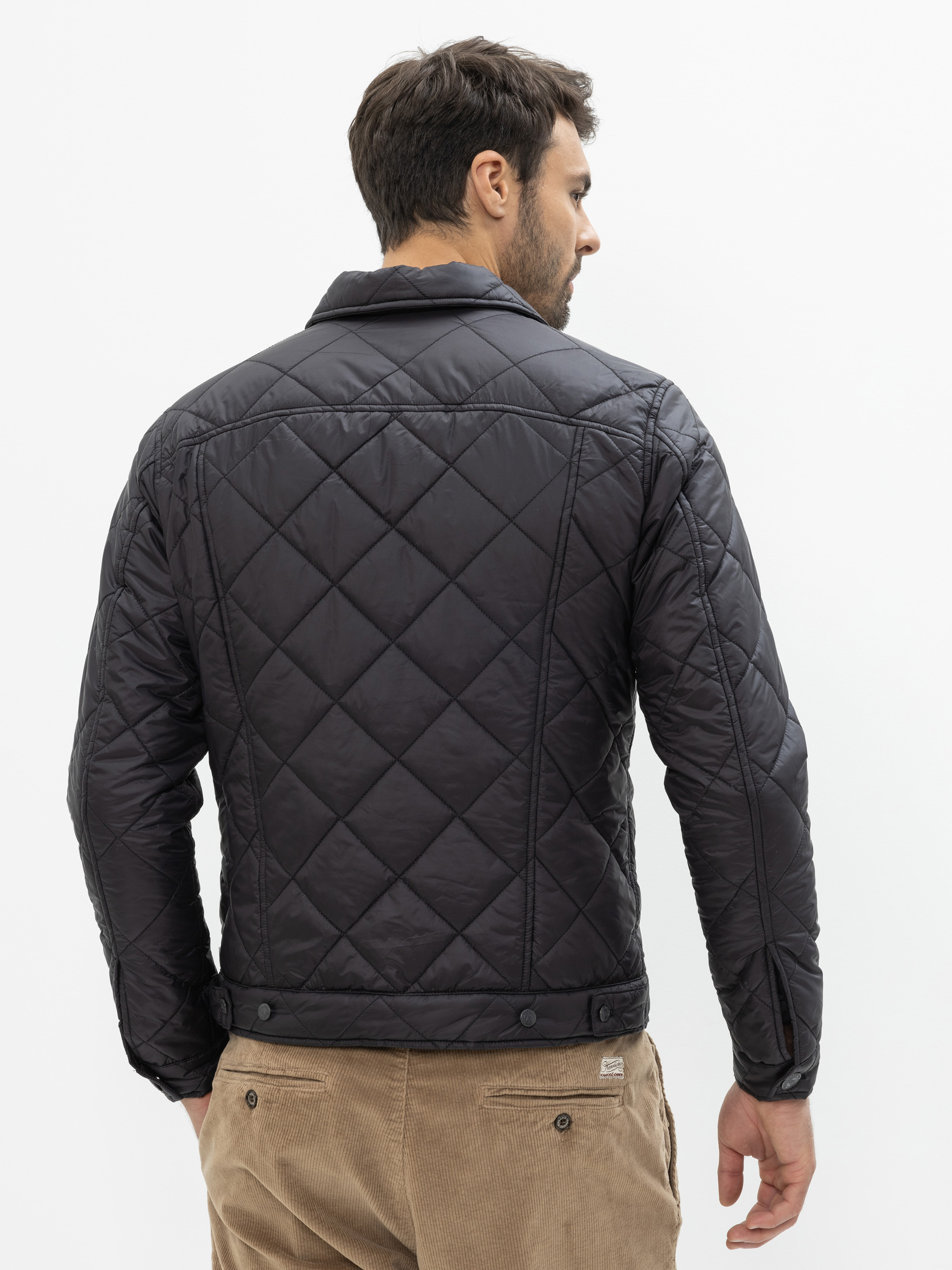 Ein Mann mit kurzen dunklen Haaren blickt von der Kamera weg und trägt eine schwarze Steppjacke und eine hellbraune Kordhose vor einem schlichten weißen Hintergrund.