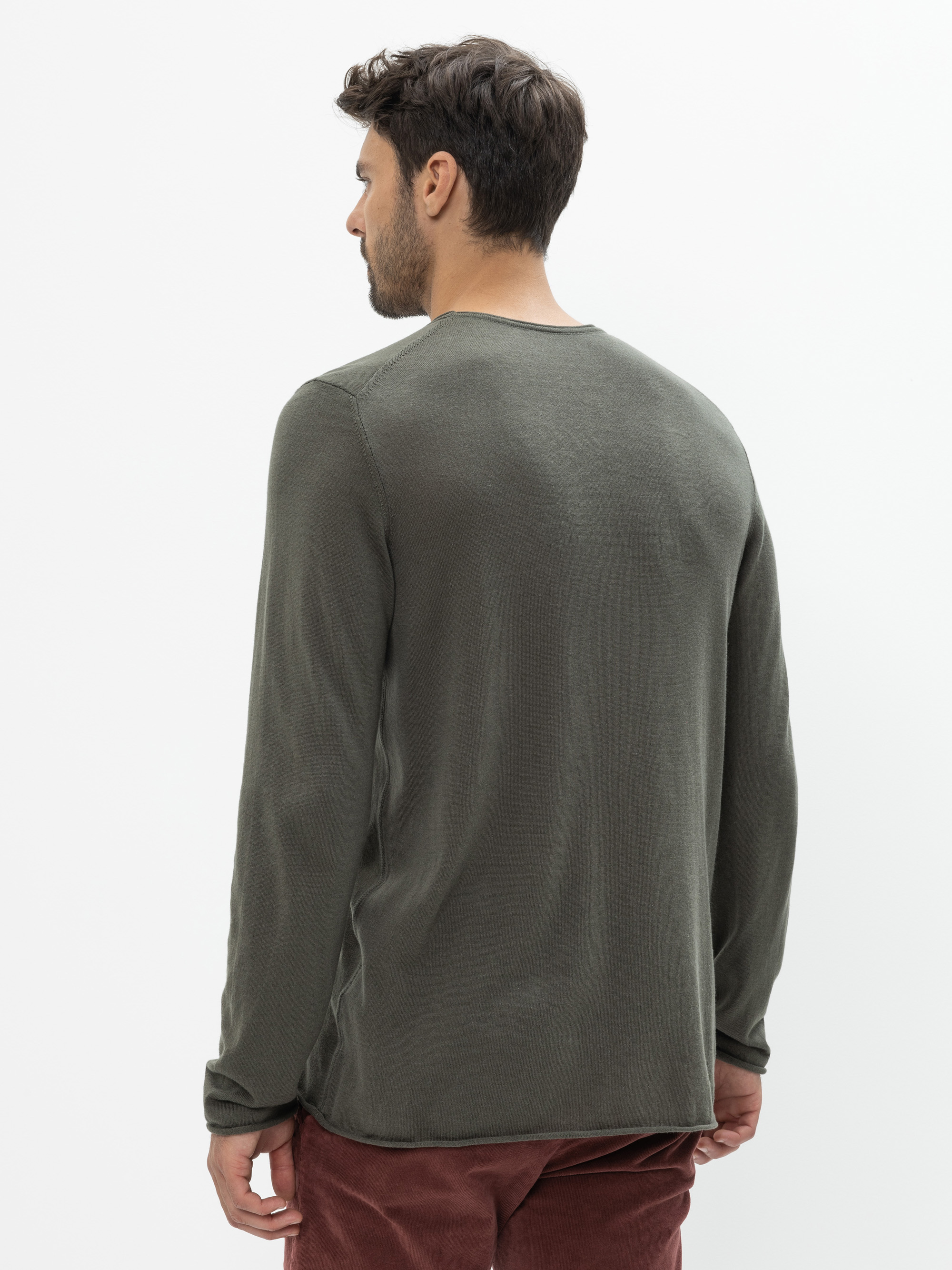 Ein Mann mit kurzen dunklen Haaren, von hinten gesehen, trägt ein langärmeliges olivgrünes Hemd und eine rötlich-braune Hose und steht vor einem schlichten weißen Hintergrund.