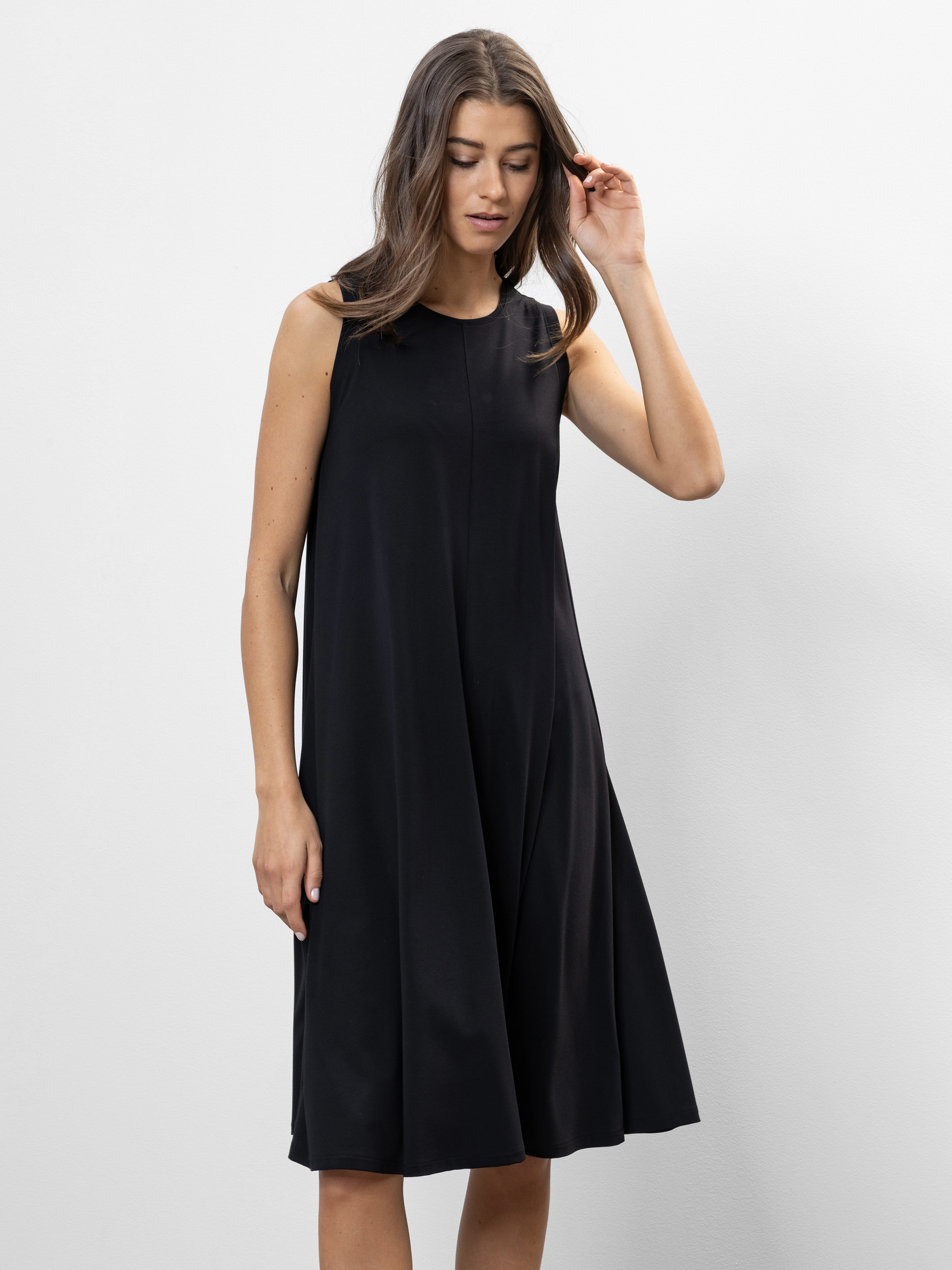 Eine Frau mit langen braunen Haaren und einem ärmellosen, knielangen schwarzen Kleid steht vor einem schlichten weißen Hintergrund und blickt nach unten, wobei sie mit einer Hand ihr Haar berührt.