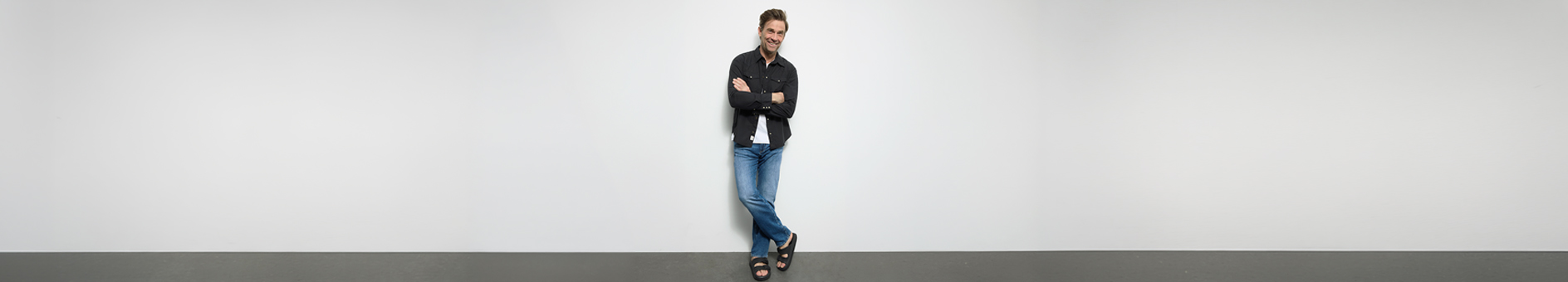 Ein Mann in einem dunklen Hemd, blauen Jeans und Sandalen steht mit verschränkten Armen an einer schlichten weißen Wand in einem fast leeren Raum.