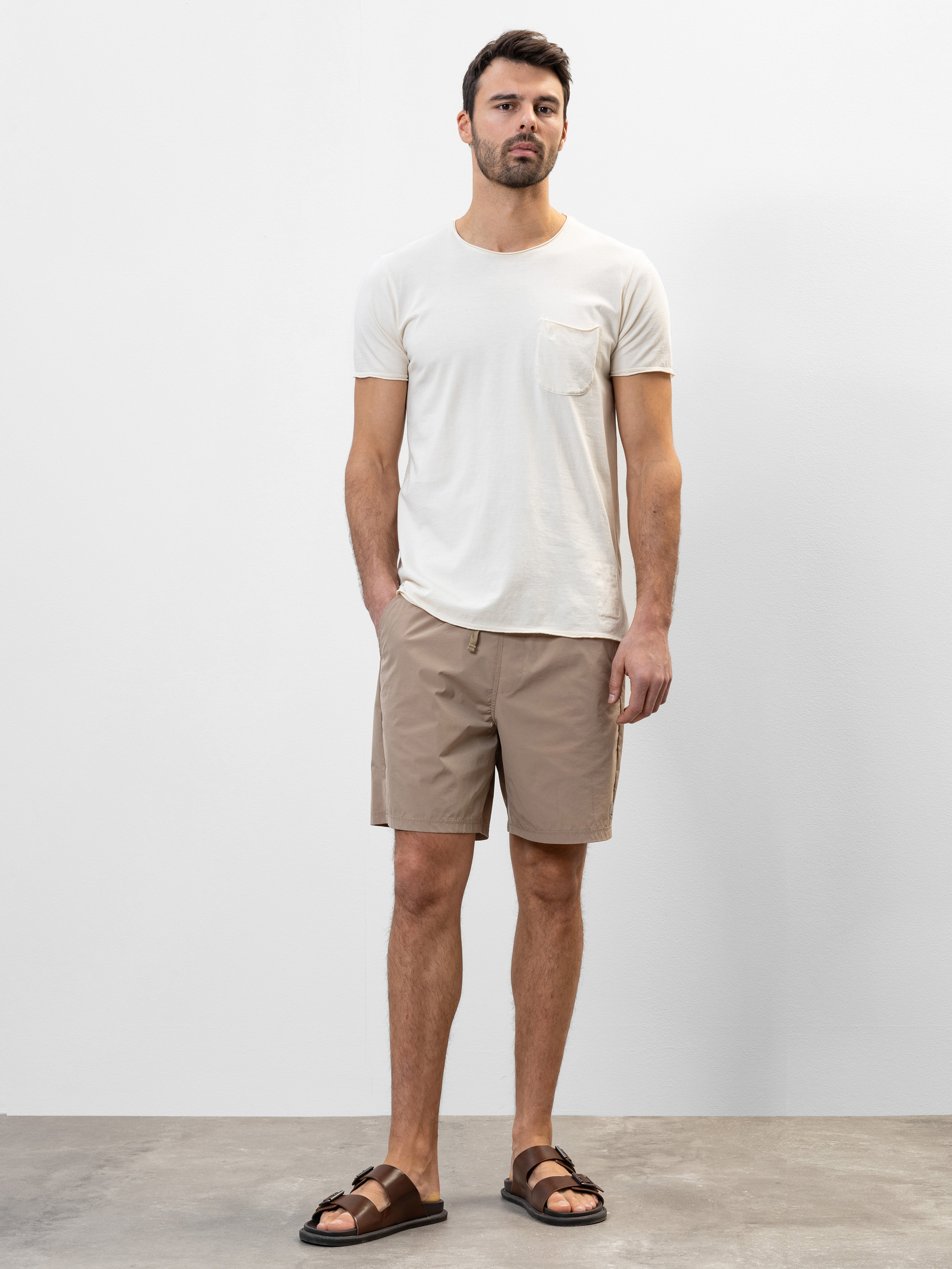 Ein Mann steht an einer schlichten weißen Wand und trägt ein weißes T-Shirt, beigefarbene Shorts und braune Sandalen. Er hat kurzes Haar und einen Bart, seine Hände liegen entspannt an den Seiten und sein Gesichtsausdruck ist neutral.