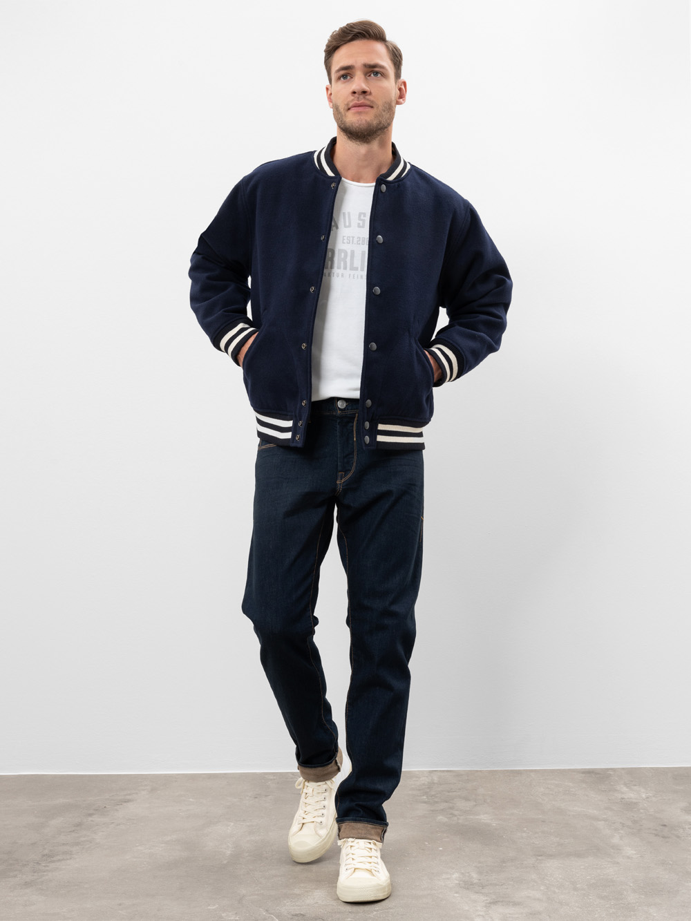 Ein Mann steht vor einem schlichten weißen Hintergrund und trägt eine marineblaue Sportjacke mit weißem Rand, ein weißes T-Shirt, dunkelblaue Jeans und weiße Turnschuhe. Er hat die Hände in den Jackentaschen und blickt leicht zur Seite.