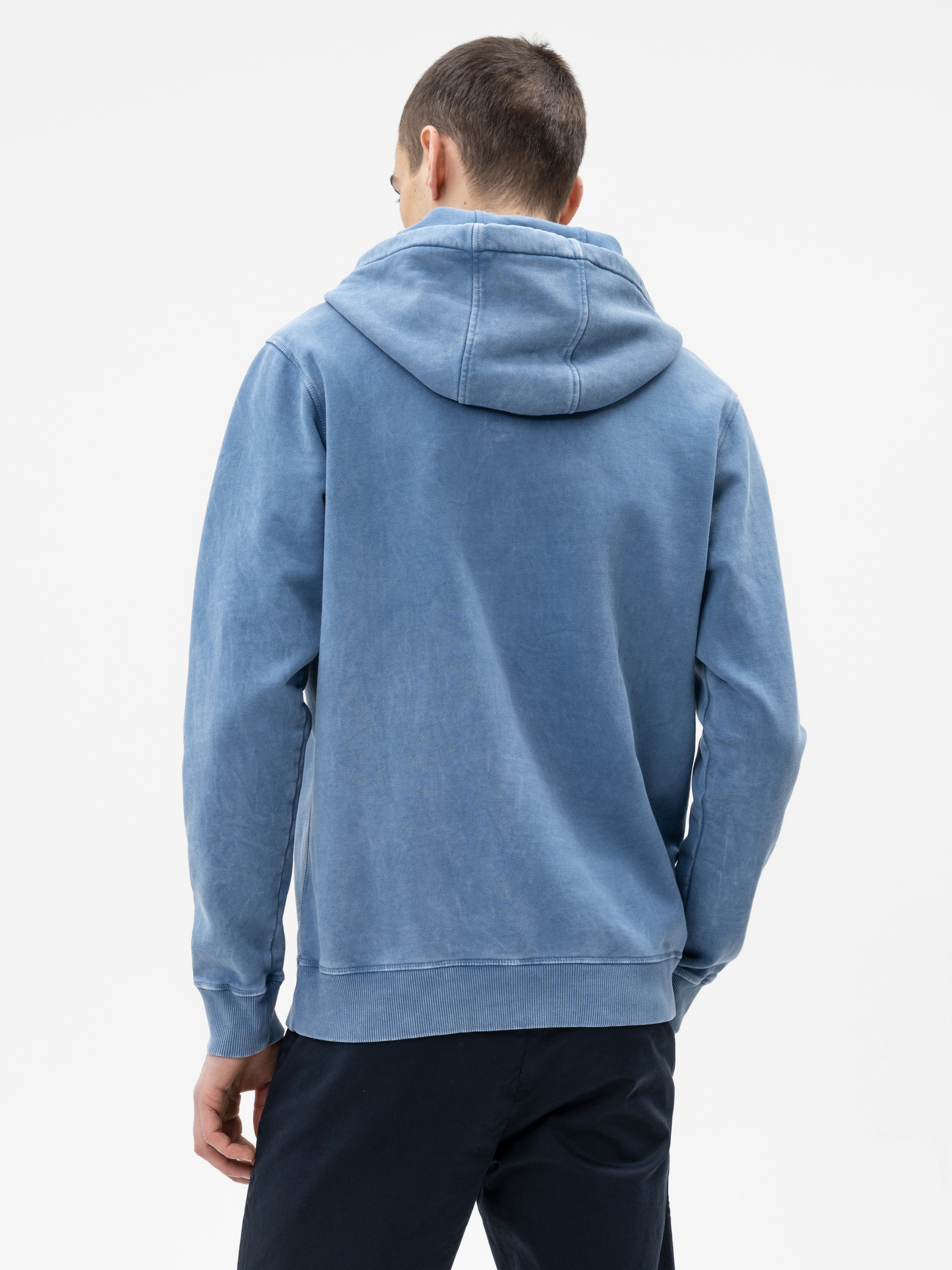 Ein Mann steht mit dem Rücken zur Kamera, trägt einen hellblauen Kapuzenpullover und eine dunkle Hose vor einem schlichten weißen Hintergrund.