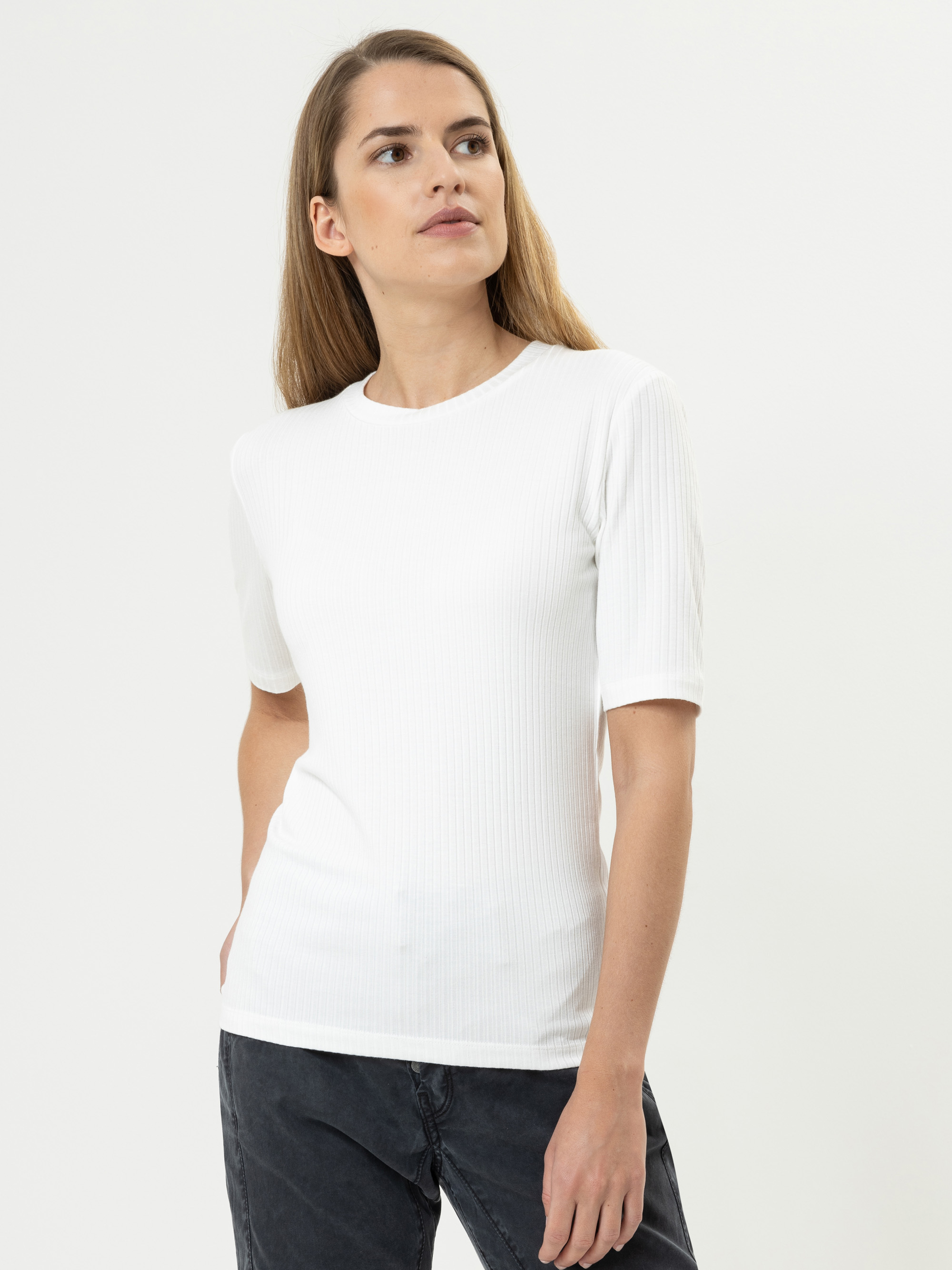 Eine Frau mit langem, hellbraunem Haar trägt ein kurzärmeliges, weißes, geripptes Oberteil und dunkle Jeans. Sie steht vor einem schlichten weißen Hintergrund und blickt leicht nach links.