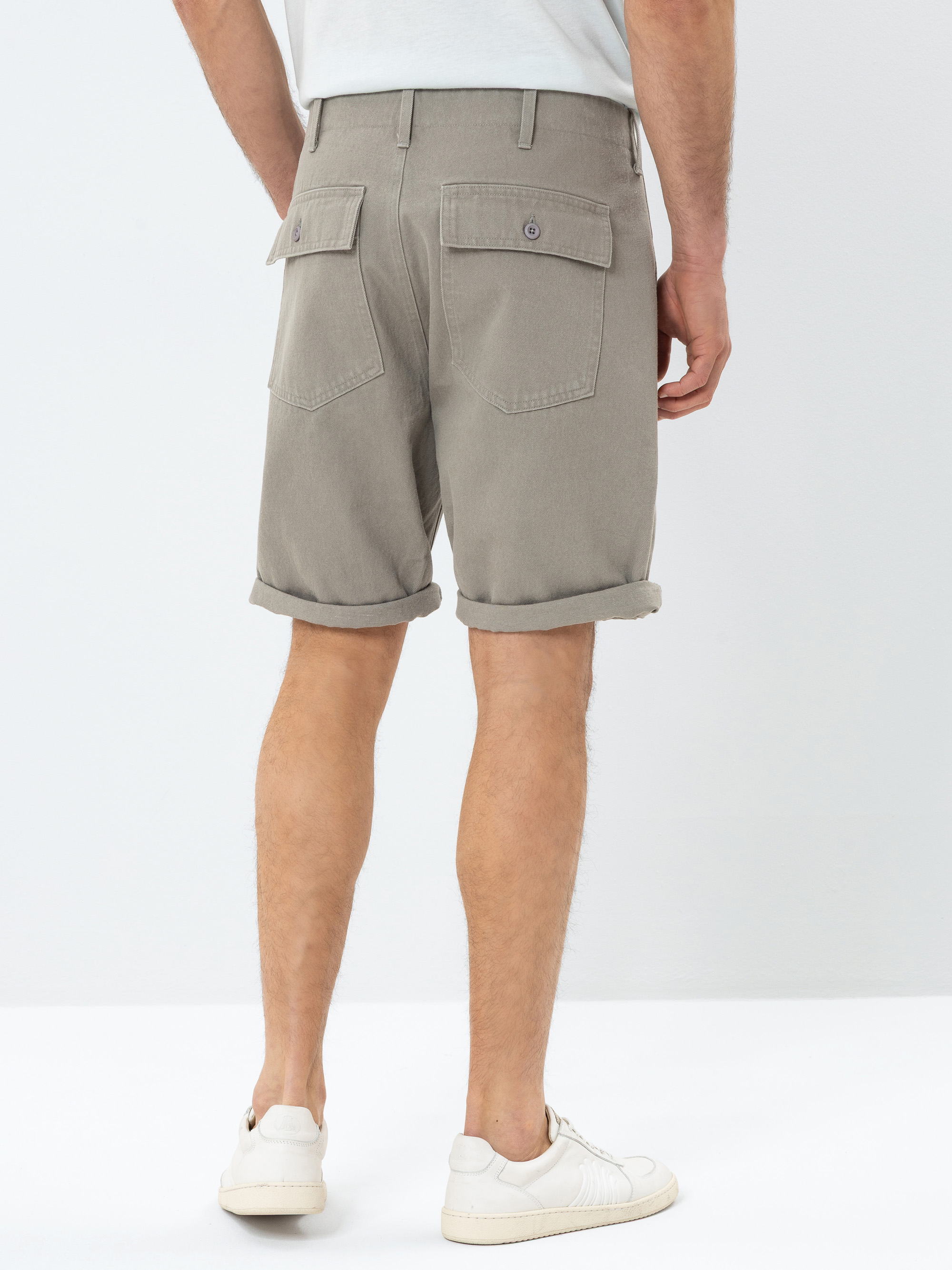 Eine Person in hellgrauen, gekrempelten Shorts mit Gesäßtaschen und weißen Turnschuhen, die vor einem schlichten, hellen Hintergrund steht, von hinten gesehen.