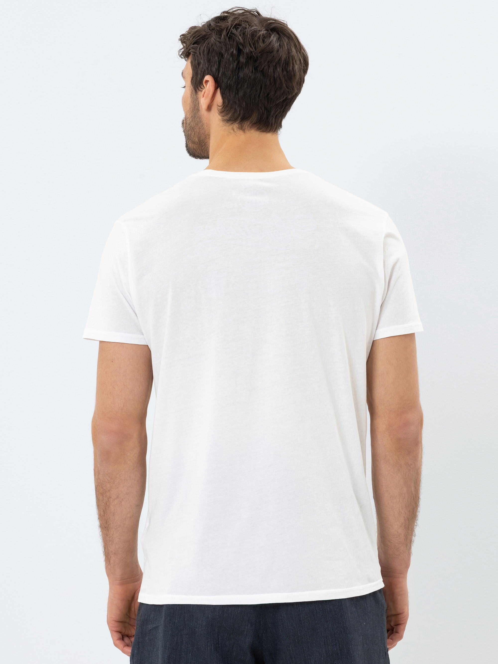 Ein Mann mit kurzen braunen Haaren wird von hinten gezeigt. Er trägt ein schlichtes weißes, kurzärmeliges T-Shirt und eine dunkle Hose und steht vor einem hellen Hintergrund.