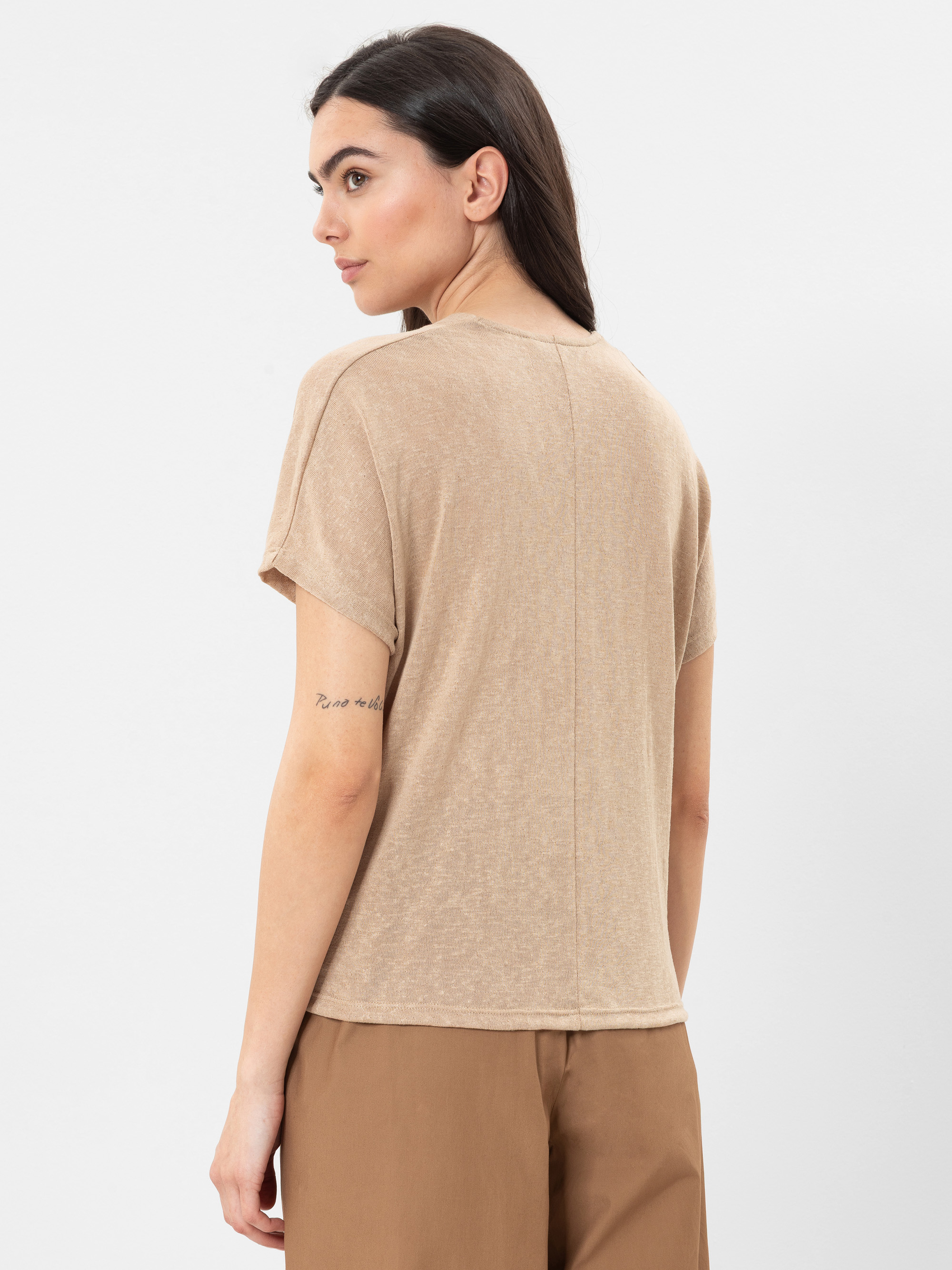 Eine Frau mit langen dunklen Haaren wird von hinten gezeigt. Sie trägt ein lockeres beiges T-Shirt und eine braune Hose. Sie hat eine sichtbare Tätowierung auf ihrem linken Unterarm und steht vor einem schlichten weißen Hintergrund.
