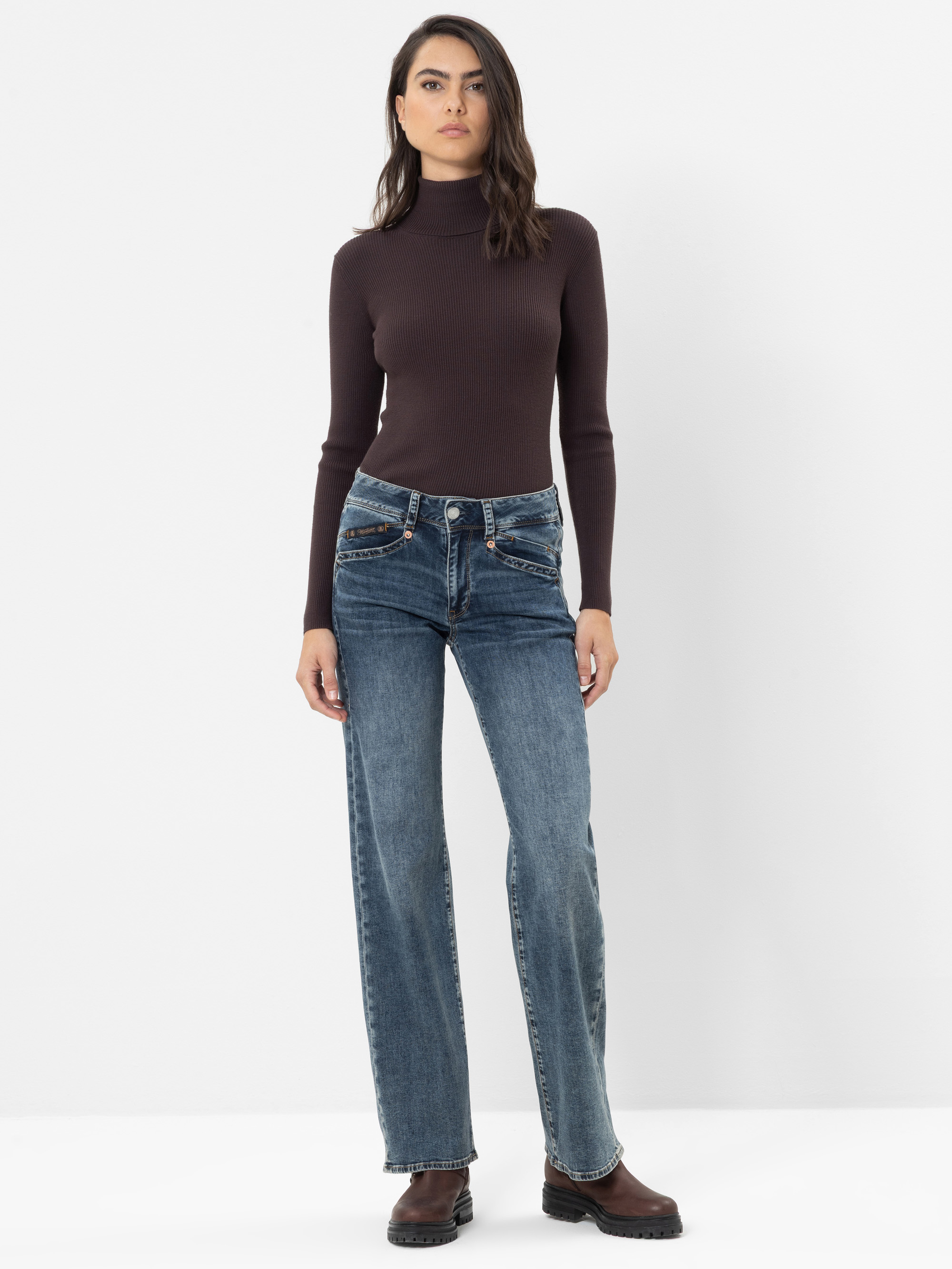 Eine Frau steht vor einem schlichten weißen Hintergrund. Sie trägt einen eng anliegenden dunkelbraunen Rollkragenpullover, eine blaue Jeans mit hoher Taille und geradem Bein sowie braune Schuhe; ihr Haar ist offen und ihr Gesichtsausdruck ist neutral.