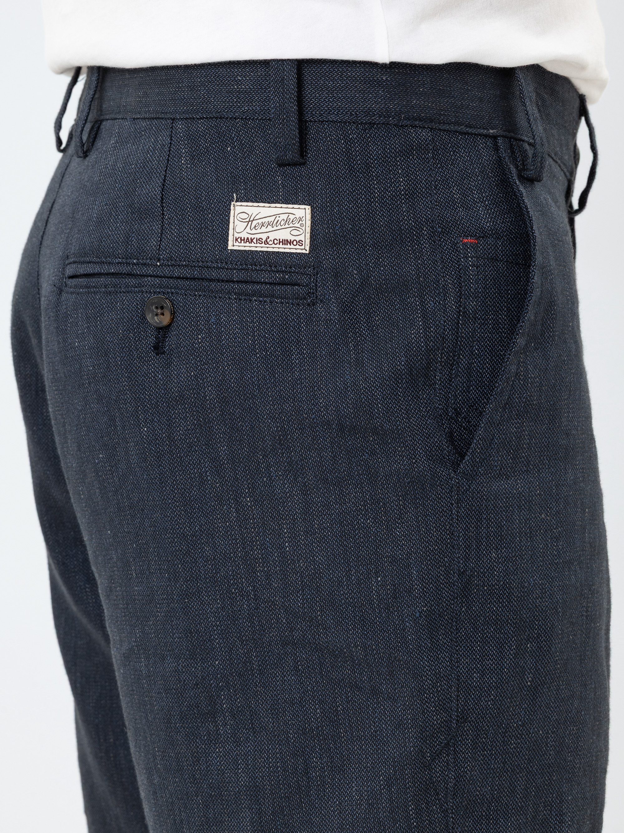 Person, die eine dunkelblaue strukturierte Hose trägt, mit einem Etikett über der Gesäßtasche, auf dem Handwerker HAKU & Co. DEUTSCHLAND. Die Hose hat Gürtelschlaufen und eine geknöpfte Gesäßtasche. Die Person trägt außerdem ein weißes Hemd.