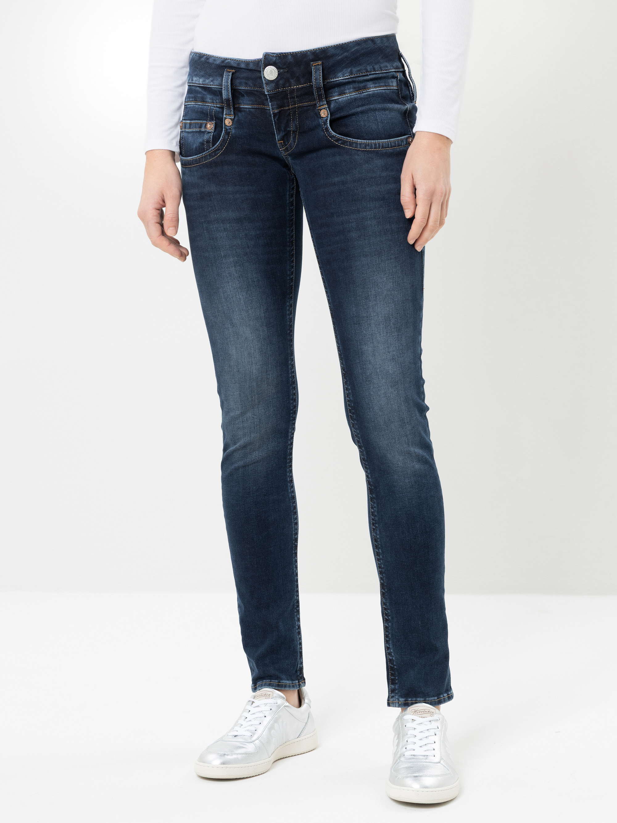 Eine Person, die eine dunkelblaue Skinny-Jeans, ein weißes Langarm-Oberteil und weiße Sneaker trägt und vor einem schlichten, hellen Hintergrund steht. Das Bild zeigt das Outfit von den Schultern bis zu den Füßen.