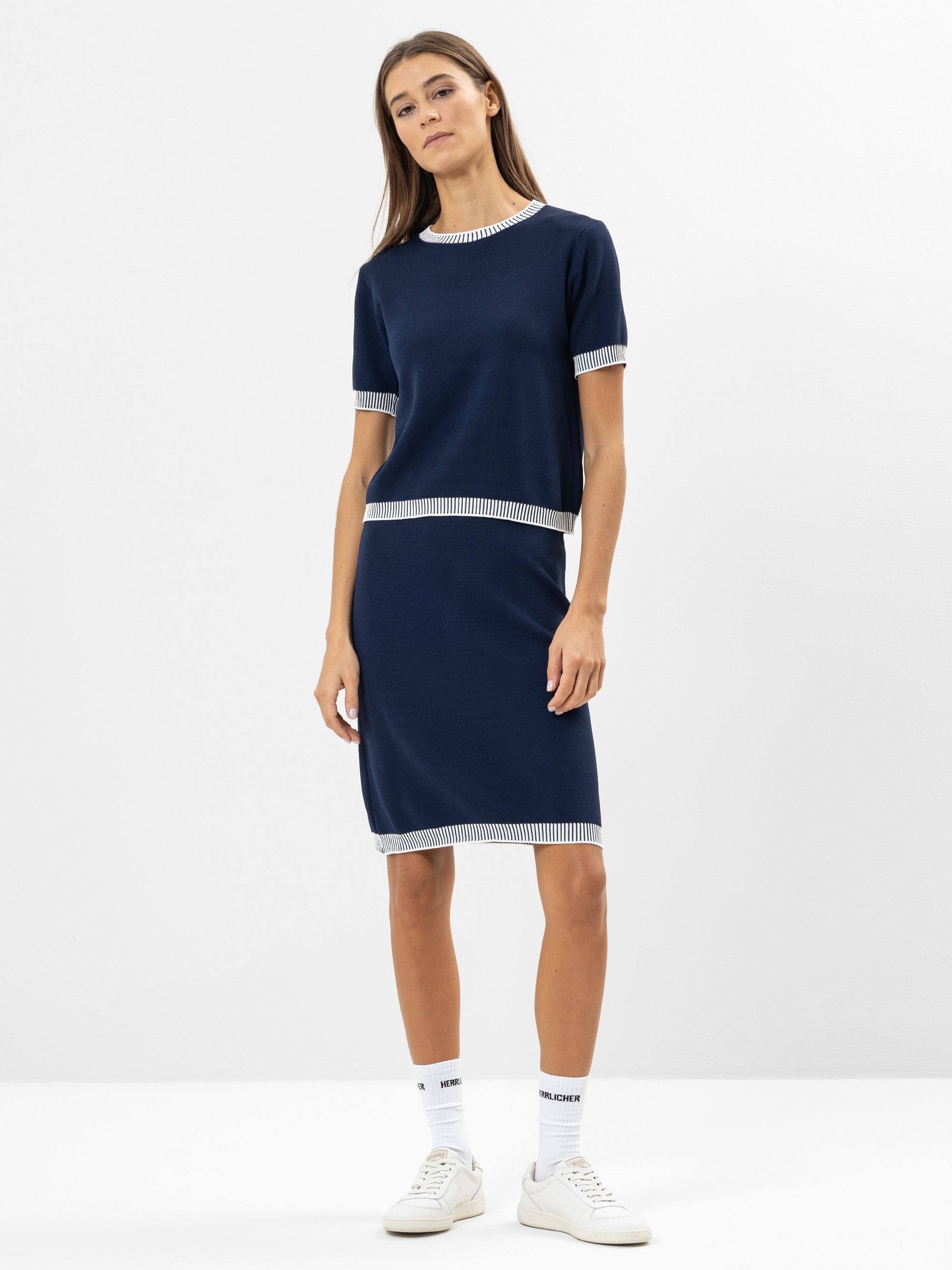 Eine Frau steht vor einem schlichten weißen Hintergrund und trägt ein marineblaues, kurzärmeliges Oberteil und einen passenden knielangen Rock mit weißem Saum, weiße Socken und weiße Turnschuhe. Sie blickt selbstbewusst in die Kamera und lässt ihre