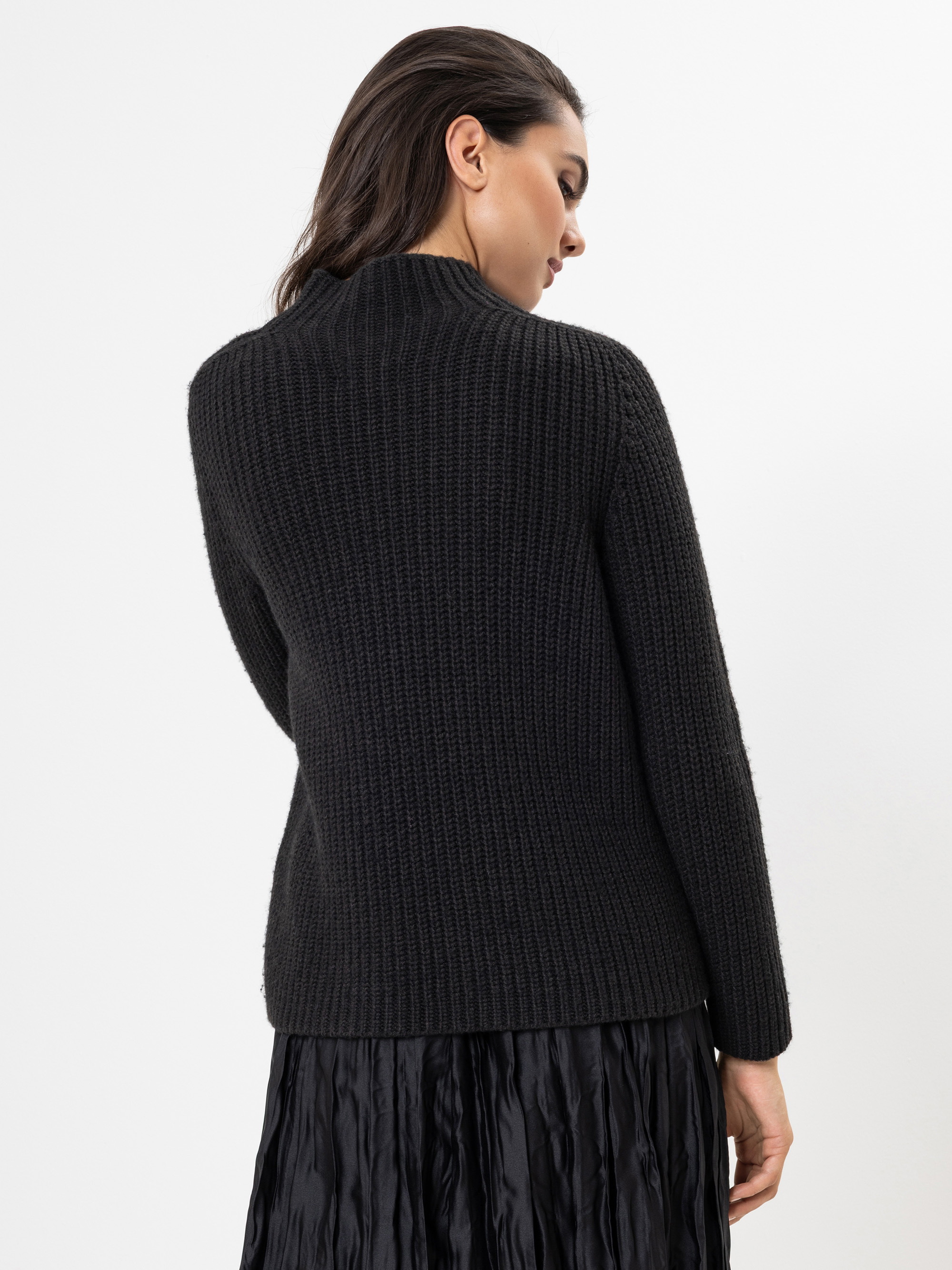 Eine Frau mit dunklem Haar steht mit dem Rücken zur Kamera und trägt einen strukturierten schwarzen Pullover und einen schwarzen Faltenrock vor einem schlichten weißen Hintergrund.