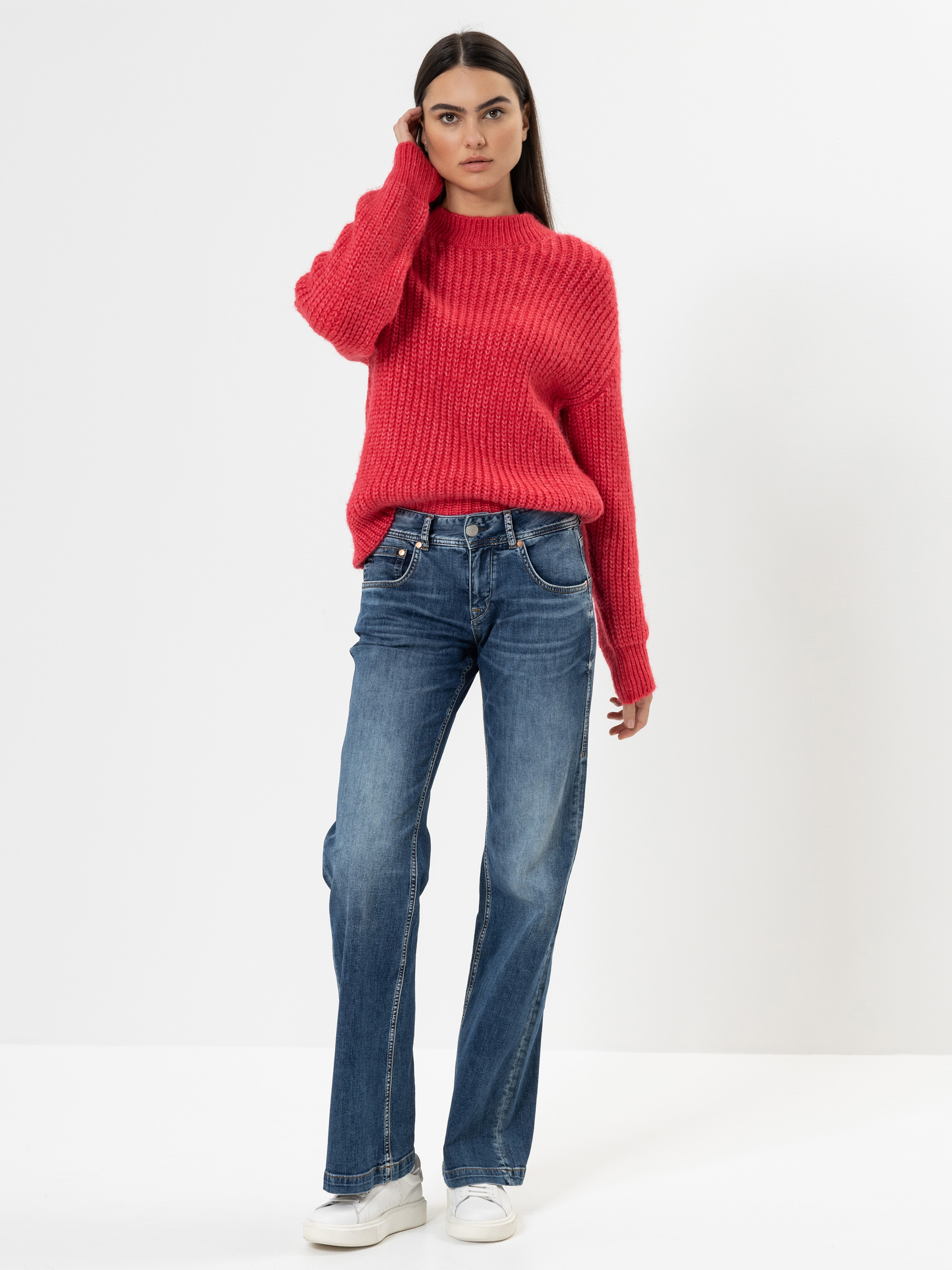 Eine Frau steht vor einem weißen Hintergrund und trägt einen roten Strickpullover, blaue Jeans und weiße Turnschuhe. Sie hat lange dunkle Haare und ihre linke Hand berührt ihren Kopf.