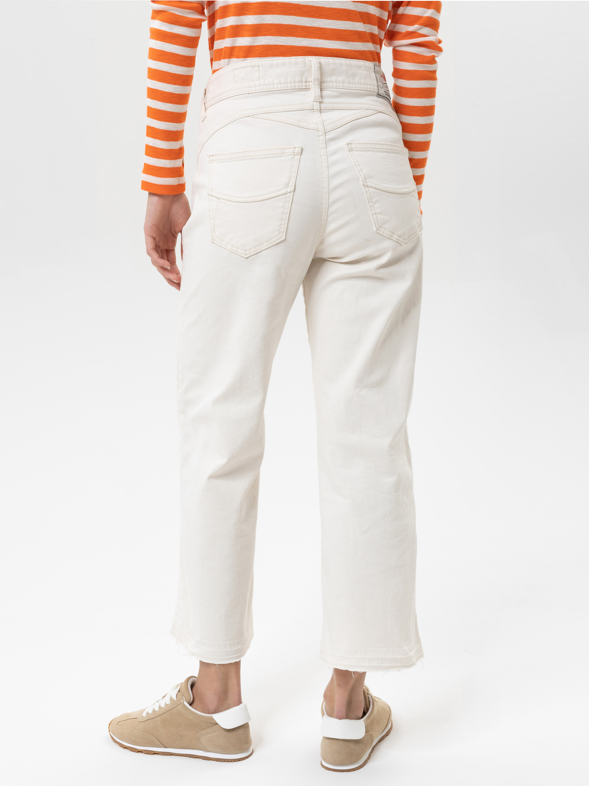 Eine Person in cremefarbenen Jeans, einem langärmeligen orange-weiß gestreiften Hemd und beigen Turnschuhen steht mit dem Rücken zur Kamera vor einem schlichten weißen Hintergrund.