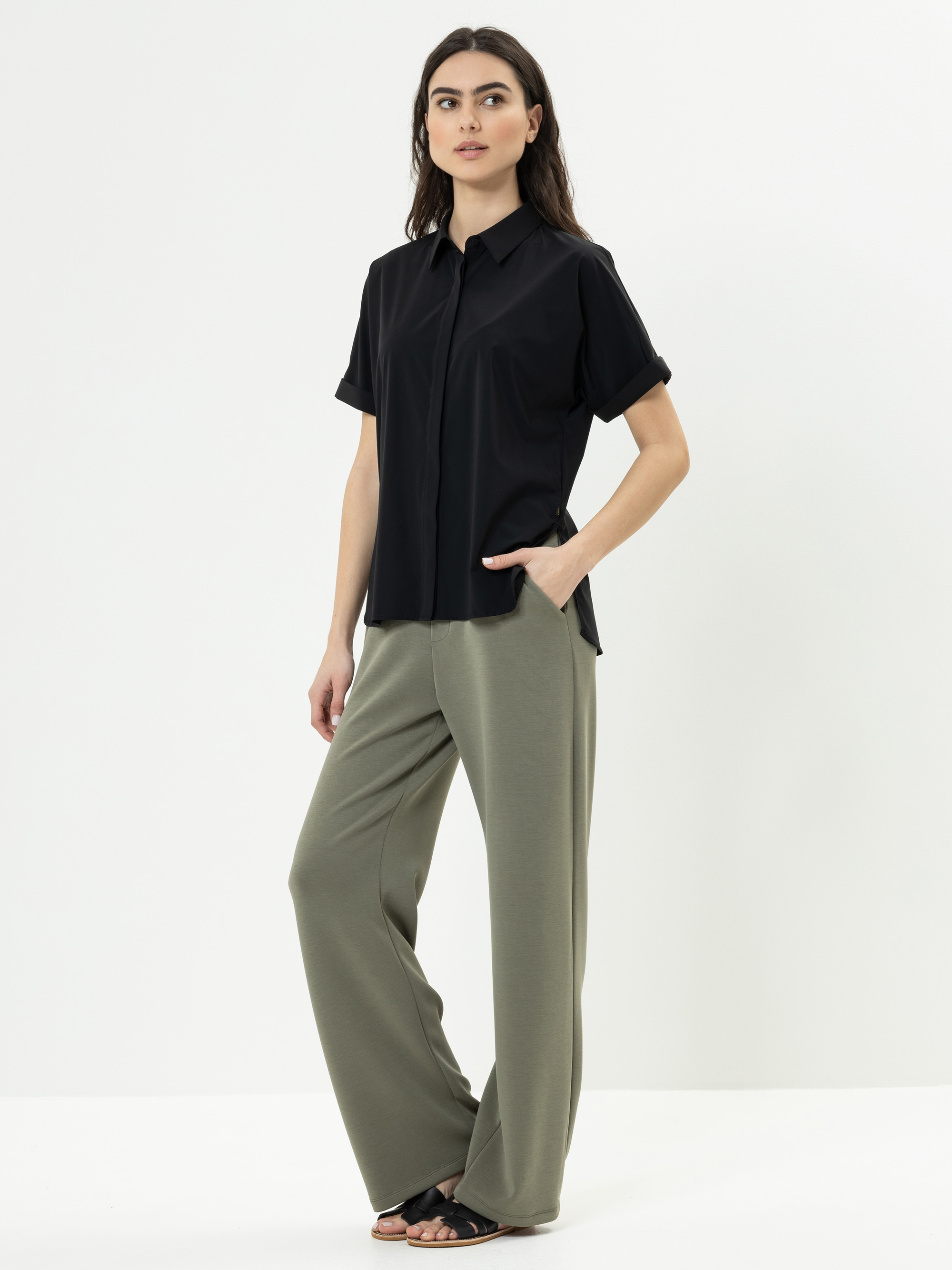 Eine Frau steht vor einem weißen Hintergrund und trägt ein kurzärmeliges schwarzes Button-up-Hemd, eine weite olivgrüne Hose und schwarze Sandalen. Sie hat eine Hand in der Tasche und blickt leicht zur Seite.