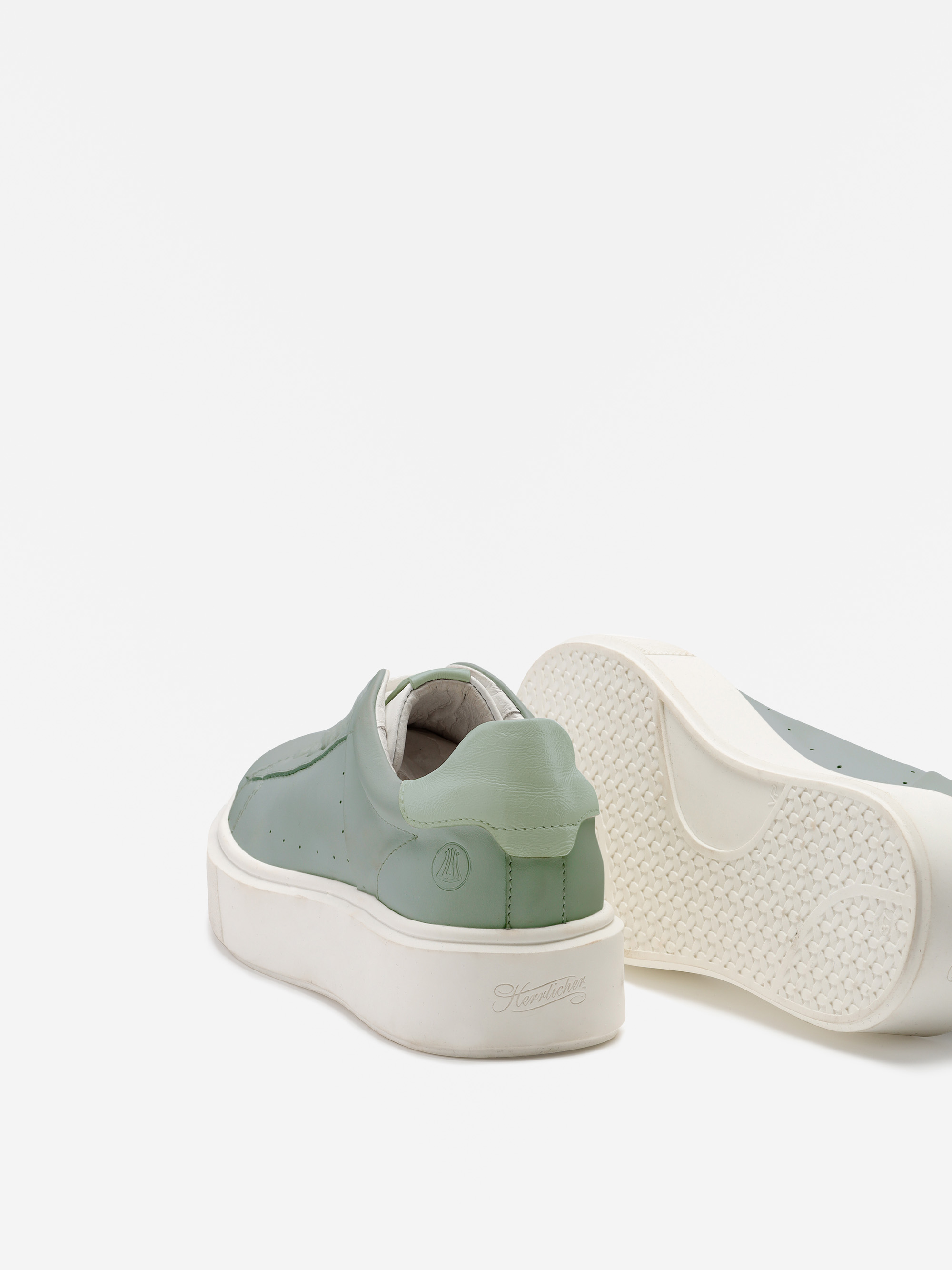 Zwei salbeigrüne Turnschuhe mit dicken weißen Sohlen sind vor einem einfarbig weißen Hintergrund abgebildet; ein Schuh steht aufrecht, während der andere auf der Seite liegt und die strukturierte Sohle zeigt.