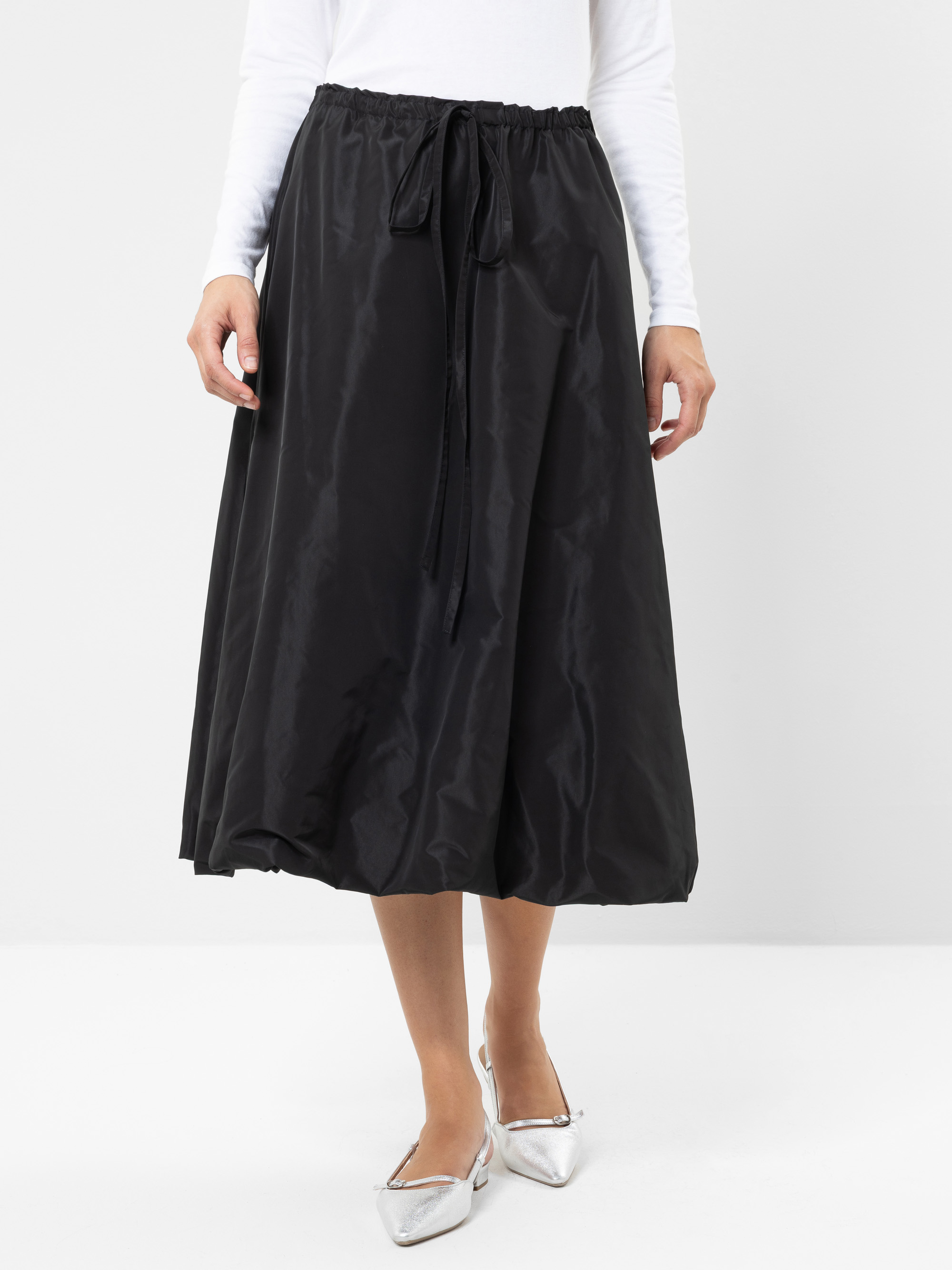 Eine Person, die ein weißes Oberteil mit langen Ärmeln, einen schwarzen Midirock mit Kordelzug in der Taille und silberne spitze Ballerinas trägt, steht vor einem schlichten weißen Hintergrund.